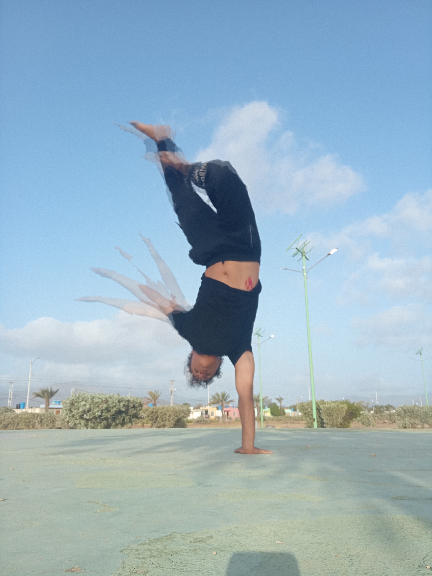 The image depicts a person performing a handstand outdoors during daylight, likely in Juan Griego, Venezuela. The subject, appearing to be male, is captured in an inverted position, balancing on one hand with their body upside down and legs bent at the knees, feet pointing towards the sky. They are wearing a black t-shirt and dark pants, and a red mark or tattoo is visible on their lower back/abdomen area.
A notable photographic effect is present, with significant motion blur around the person's limbs, creating multiple "ghosted" images, suggesting either a dynamic movement during the handstand or a multi-exposure technique to convey motion.
The setting features a flat, light green or teal paved surface in the foreground, possibly a sports court. The background consists of a clear blue sky with scattered white clouds. Several tall, slender green metal poles, which appear to be streetlights, are visible. Green bushes and some palm trees are also present in the mid-ground, with distant buildings and structures on the horizon, consistent with a semi-urban or coastal environment. The bright natural light indicates it is daytime, likely morning or afternoon. No visible text is present.
