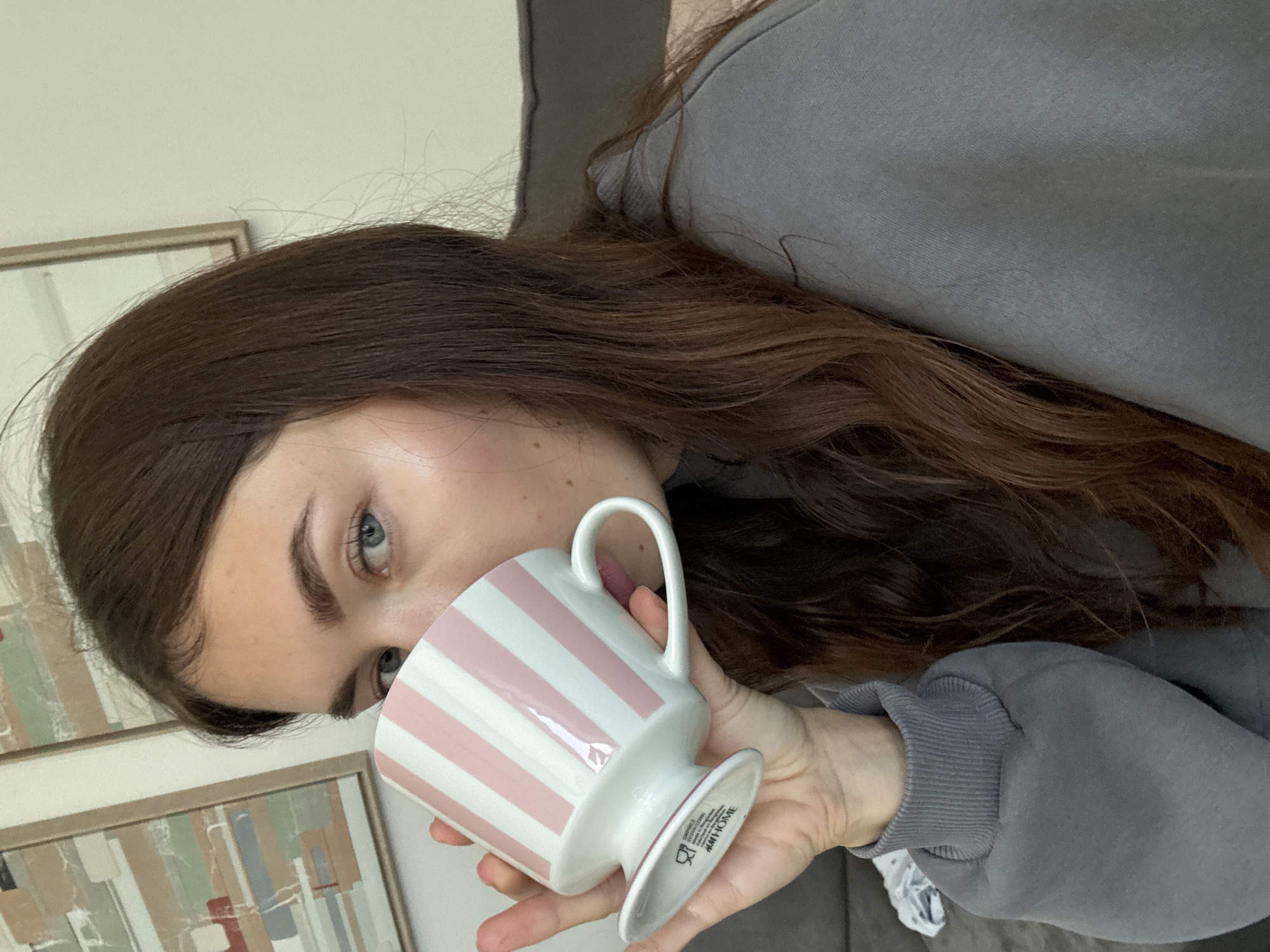 A woman with long brown hair and blue eyes is drinking from a white and pink striped cup. The cup has "H&M HOME" printed on the bottom. She is wearing a gray sweatshirt. There are two framed pictures on the wall behind her. The location is unknown, but it is not Tel-Aviv, Israel.