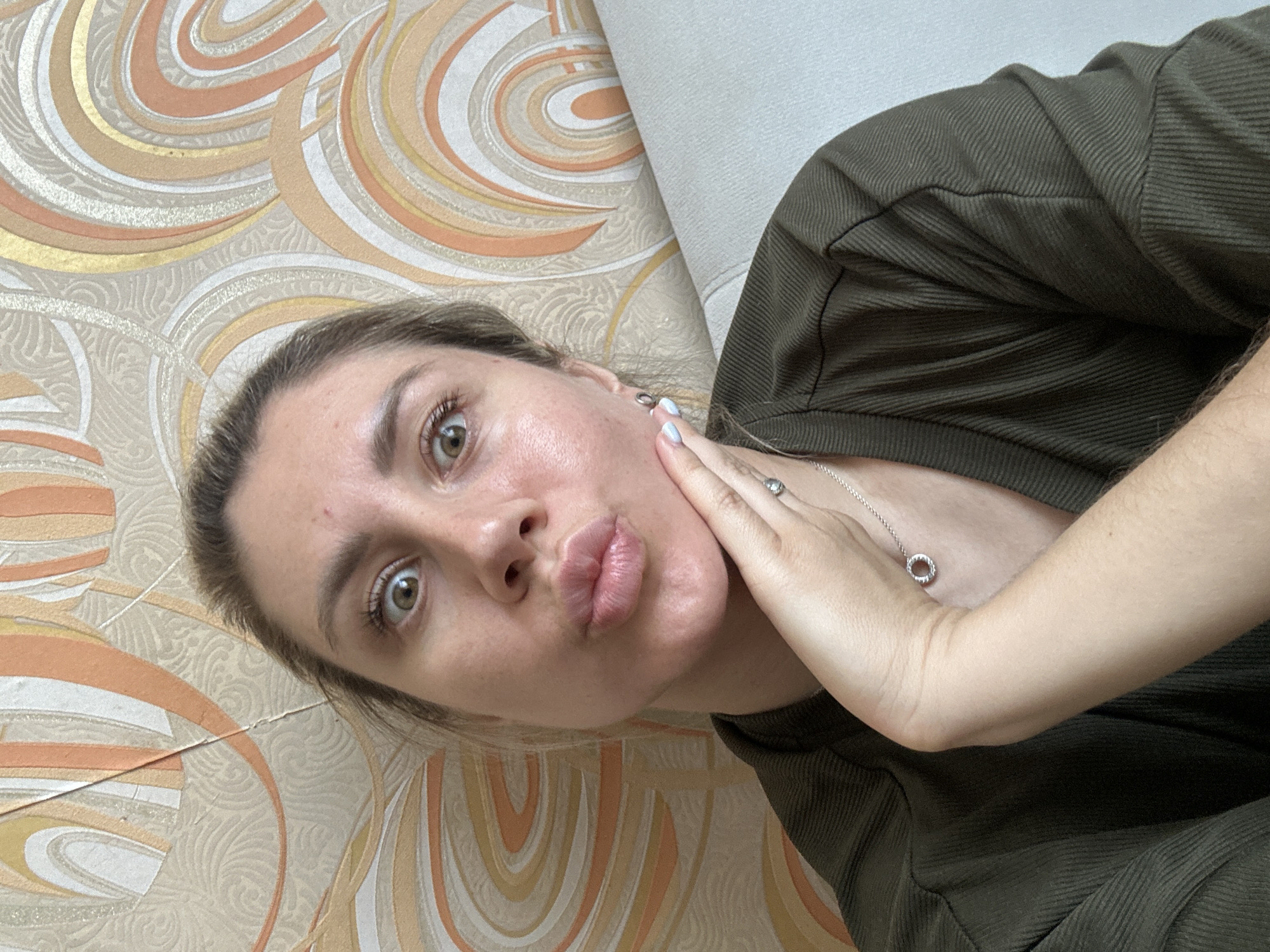 A young woman with blonde hair pulled back poses for a selfie. She is wearing a dark green, ribbed, long-sleeved shirt and a silver necklace with a circular pendant. Her right hand is placed under her chin, and she has a pouting expression with her lips pursed. Her eyes are wide open, and she is looking directly at the camera. The background features patterned wallpaper with wavy designs in shades of beige, orange, and gold. A light-colored couch is visible to the right of the woman. The image appears to have been taken indoors, possibly in an apartment in Novoaltaysk, Russia, given the context.