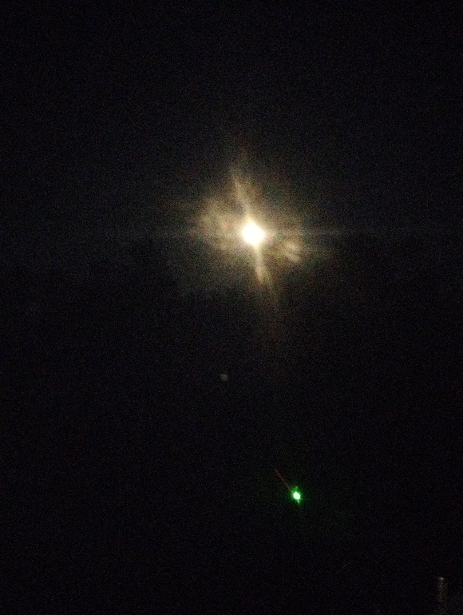 The image depicts a dark night sky with a bright, star-like object in the upper center, likely the moon or a bright star, with light rays emanating from it. In the lower right quadrant, there is a small, bright green light source with a faint red trail, possibly a laser pointer or a distant aircraft light. The background is predominantly black, indicating nighttime. No specific location in the city of Russkii, Russia can be determined from the image. There are no discernible people, specific buildings, or other urban environmental features.