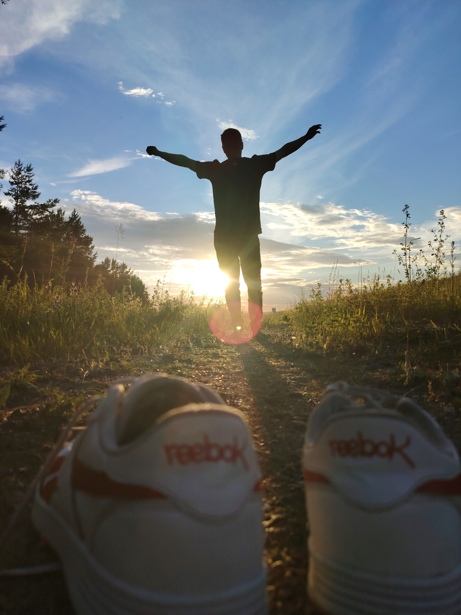 The image depicts a low-angle shot of a person silhouetted against a sunset sky. The individual stands with arms outstretched, facing away from the camera, with the sun directly behind them. In the foreground, a pair of white Reebok sneakers with red accents are blurred and out of focus. The setting appears to be a rural or natural environment, with tall grass and trees visible in the background, illuminated by the warm light of the setting sun. The sky is a mix of blue and white clouds, with sunbeams creating a lens flare effect. There is no specific indication of the location being the city of Chaikovsky, Russia, as the image itself does not contain any identifying landmarks or signs of an urban environment.