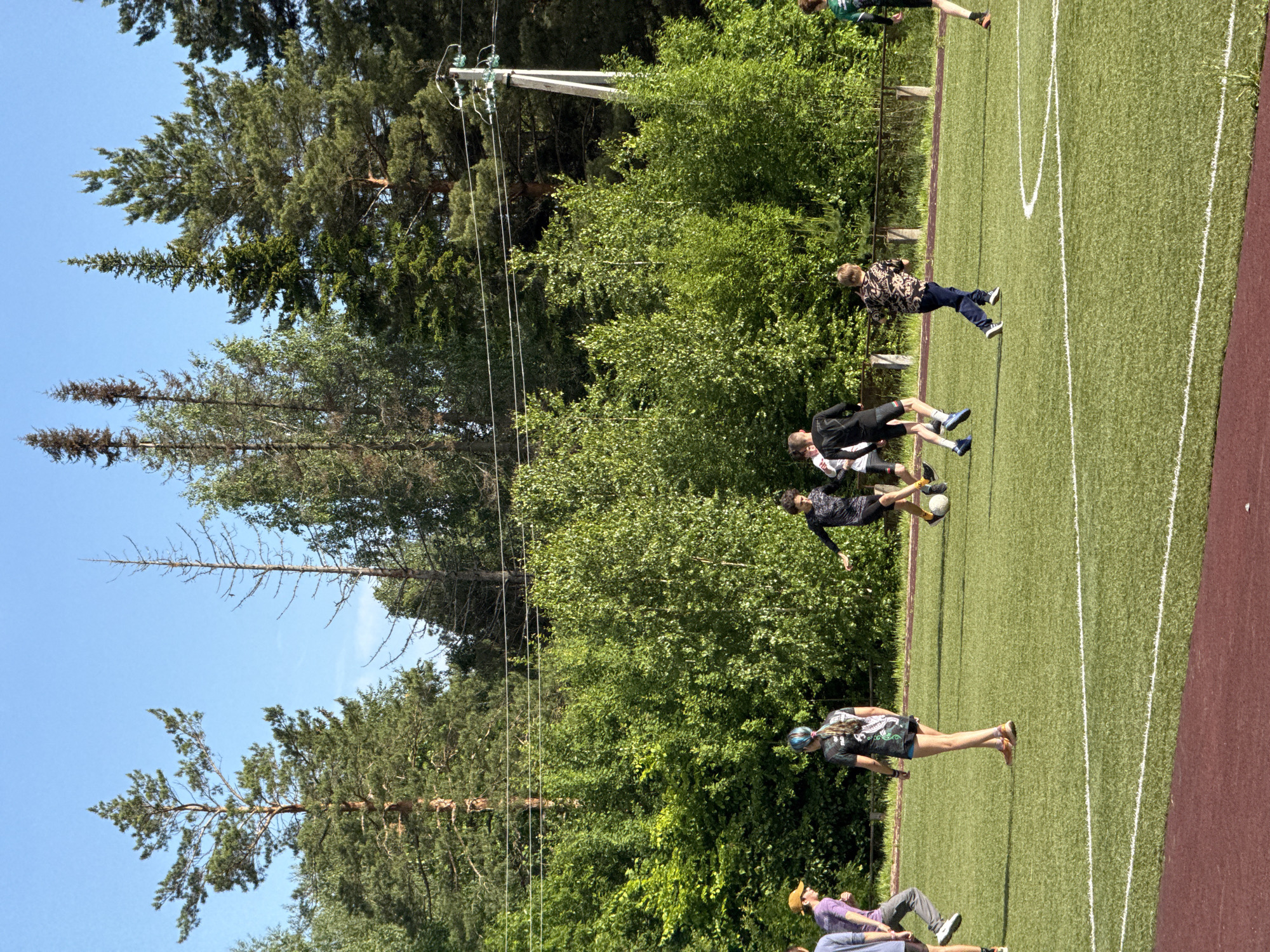The image captures an outdoor sports field under a clear, bright blue sky. The field itself is a vibrant green, marked with distinct white lines, indicating it's likely used for soccer or general athletics. A reddish-brown track or border runs along the right edge of the playing area.
A dense line of trees, including tall pines and deciduous trees, forms a lush green background across the upper half of the image. On the left side, a utility pole with multiple electrical wires and insulators rises prominently from behind the treeline.
Several individuals are present on and around the field. In the foreground, towards the left, two people are partially visible; one wears a purple shirt, grey pants, and a yellow cap, while another is in a blue t-shirt. Further back, a person in a black and green patterned t-shirt, shorts, and a light blue helmet or headband stands on the field. Near the center, two individuals are actively engaged with a white soccer ball; one in a black long-sleeved shirt and shorts appears to be kicking it. To their right, another person in a black and white patterned shirt and dark pants walks or jogs across the field. In the far background, on the right, a person in a green and black top is seen moving off the field.
The activity appears to be a casual sports gathering or practice session, possibly soccer, given the presence of the ball and the field markings. The bright sunlight and clear sky indicate a sunny day with pleasant weather. The setting is a recreational outdoor space, likely a community field or school sports ground, bordering a wooded area. No visible text is present in the image. The location context provided is Прикамский, Russia.