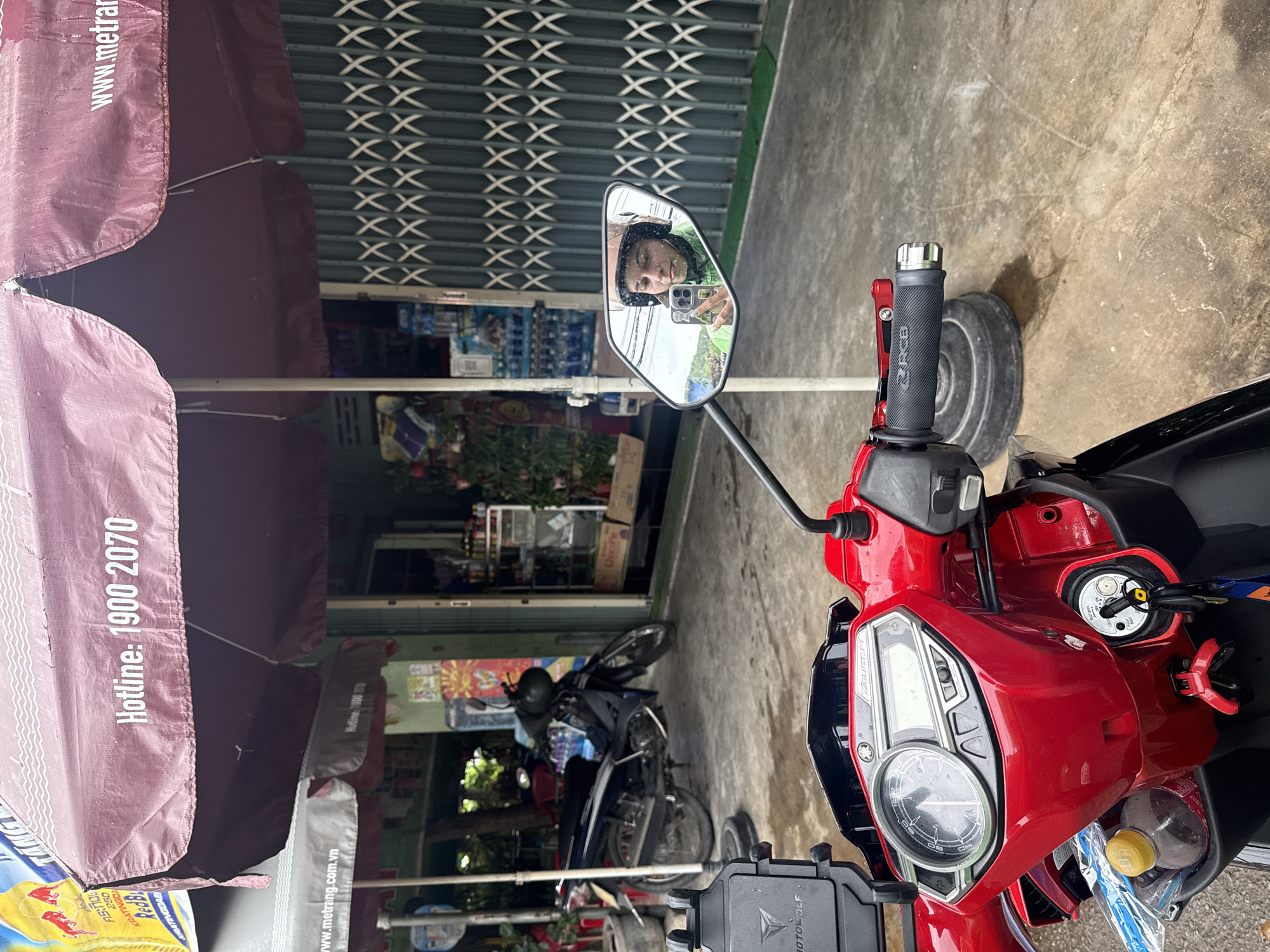 The image captures a street scene in Nha Trang, Vietnam, from the perspective of someone sitting on a red scooter. In the foreground, the handlebars and instrument panel of the scooter are visible, along with a bottle of drink in a storage compartment. A large, maroon umbrella providing shade dominates the upper portion of the image, with "Hotline: 1900 2070" printed on it. To the left, a smaller grey umbrella displays the text "www.metrang.com.vn". In the background, a dark-colored scooter is parked outside a shop with a green, collapsible metal gate. A reflection in the scooter's mirror shows the rider, wearing a helmet and a green jacket, taking a selfie with their phone. The ground appears wet, suggesting recent rain.