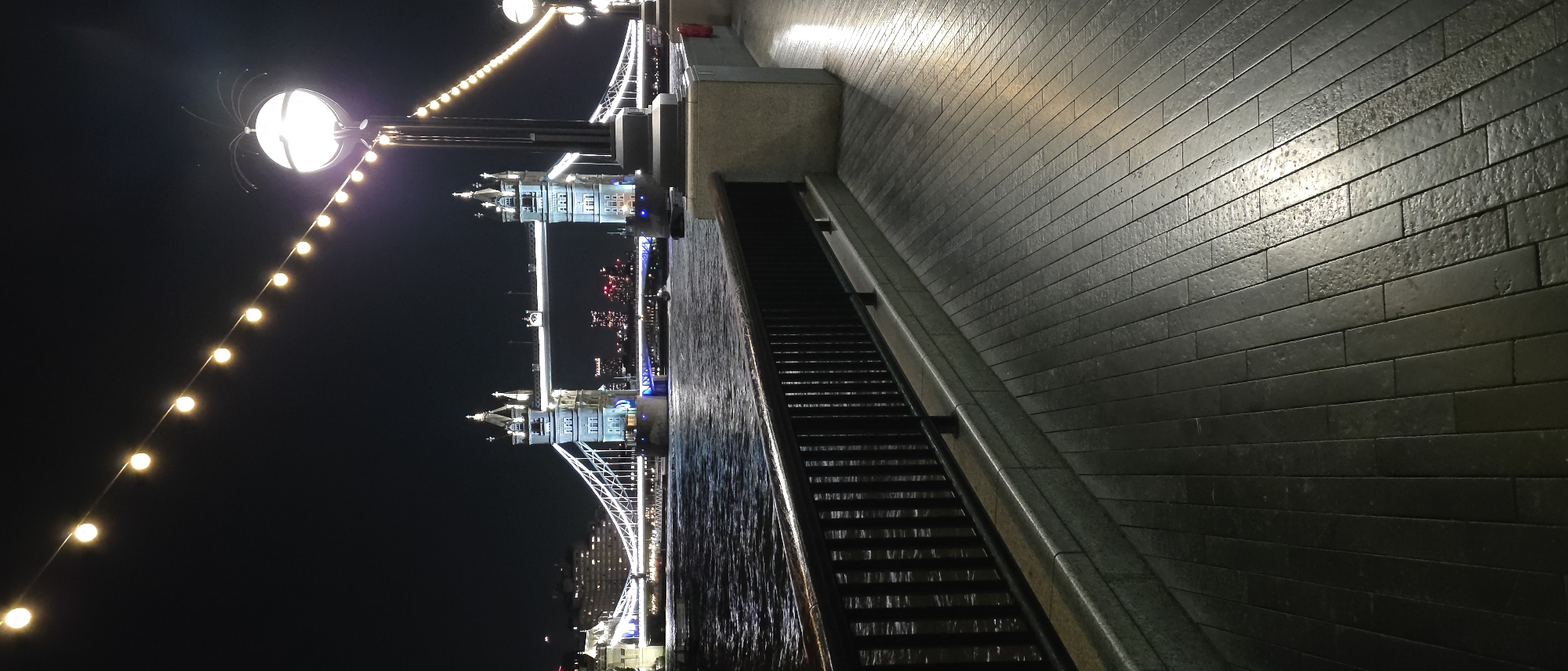 A nighttime photograph captures a section of the River Thames in London, United Kingdom, with the iconic Tower Bridge illuminated in the background. The foreground features a paved walkway with a black railing along the river's edge. String lights with glowing bulbs are visible overhead, leading the eye towards the bridge. The water of the Thames reflects the city lights and the bridge's illumination. Tower Bridge stands majestically, its towers lit with a blue hue and white accents, with its suspension elements and walkway clearly visible. The overall scene conveys a sense of urban ambiance at night.