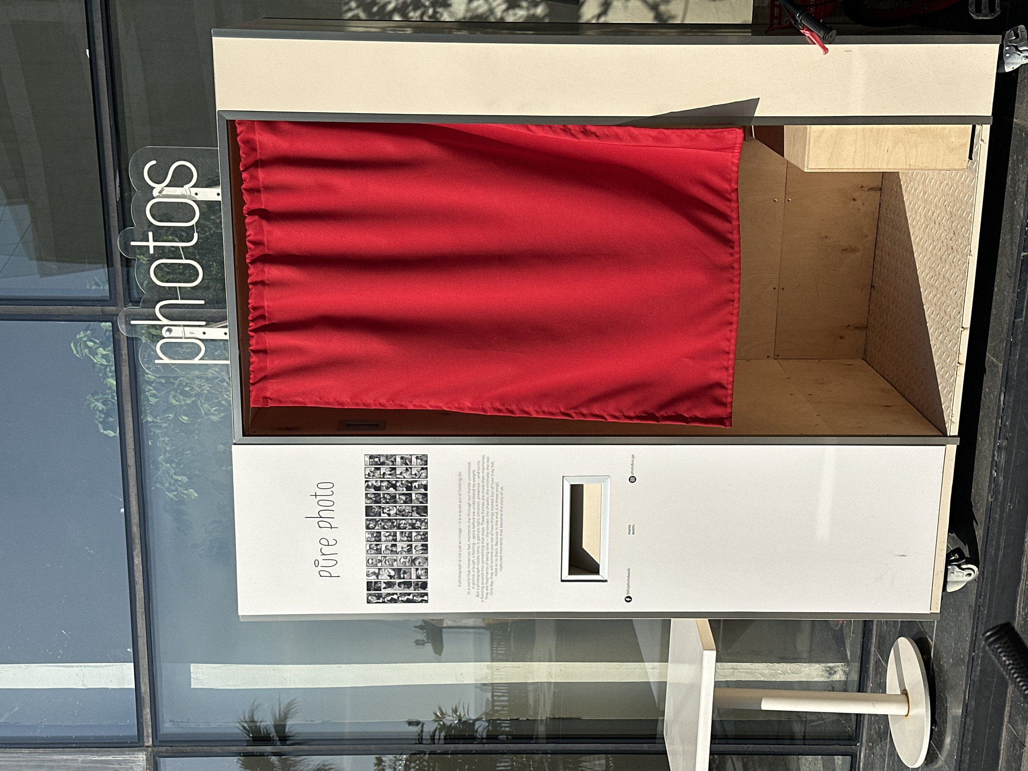 The image captures a photo booth in Batumi, Georgia. The booth, labelled "pure photo", stands against a glass front, reflecting the external environment. A red curtain acts as a backdrop inside. The booth's exterior features a panel displaying images and text, emphasizing photography's role in preserving moments. The environment includes a window with plants. A table is attached to the photo booth.