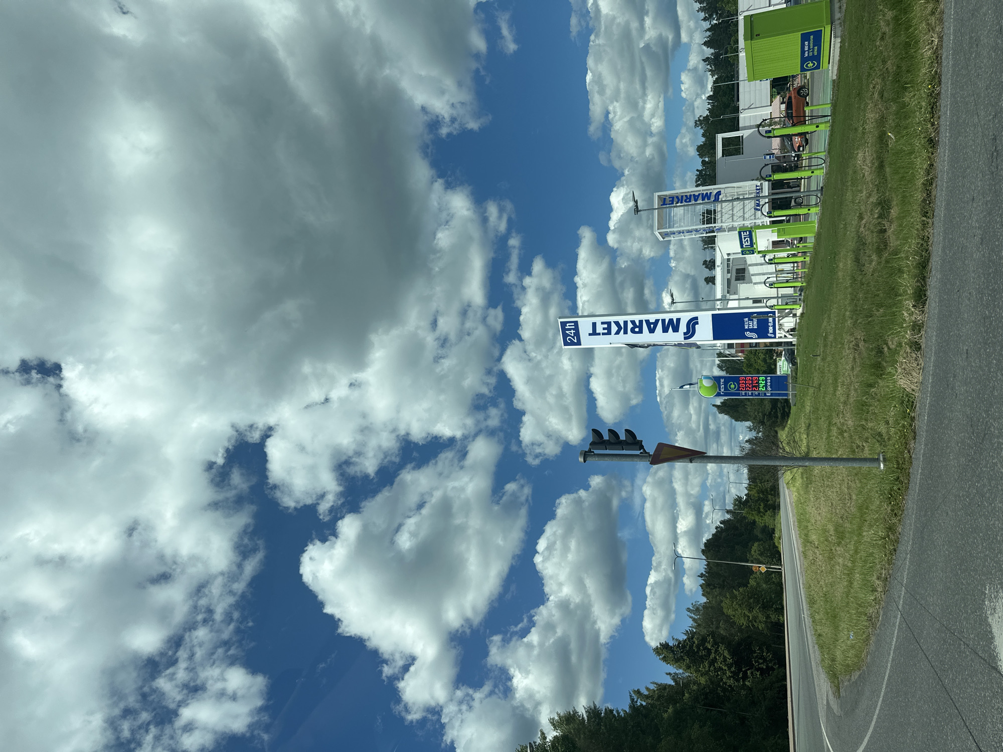 This is a street-level, eye-level, outdoor, medium shot of a gas station and convenience store under a partly cloudy blue sky. The gas station is on the right side of the image and appears to be branded with the logos "24h MARKET" and "NESTE". There are several green gas pumps visible, and a large green box on the far right with a blue sign. Behind the pumps is a white building with a modern design. A few cars are parked at the station. In the foreground, there is a paved road and a grassy area. On the left side of the image, there is a traffic light and a sign with a red triangle. A dense line of green trees borders the left side of the road. The overall scene suggests a sunny day with scattered clouds.