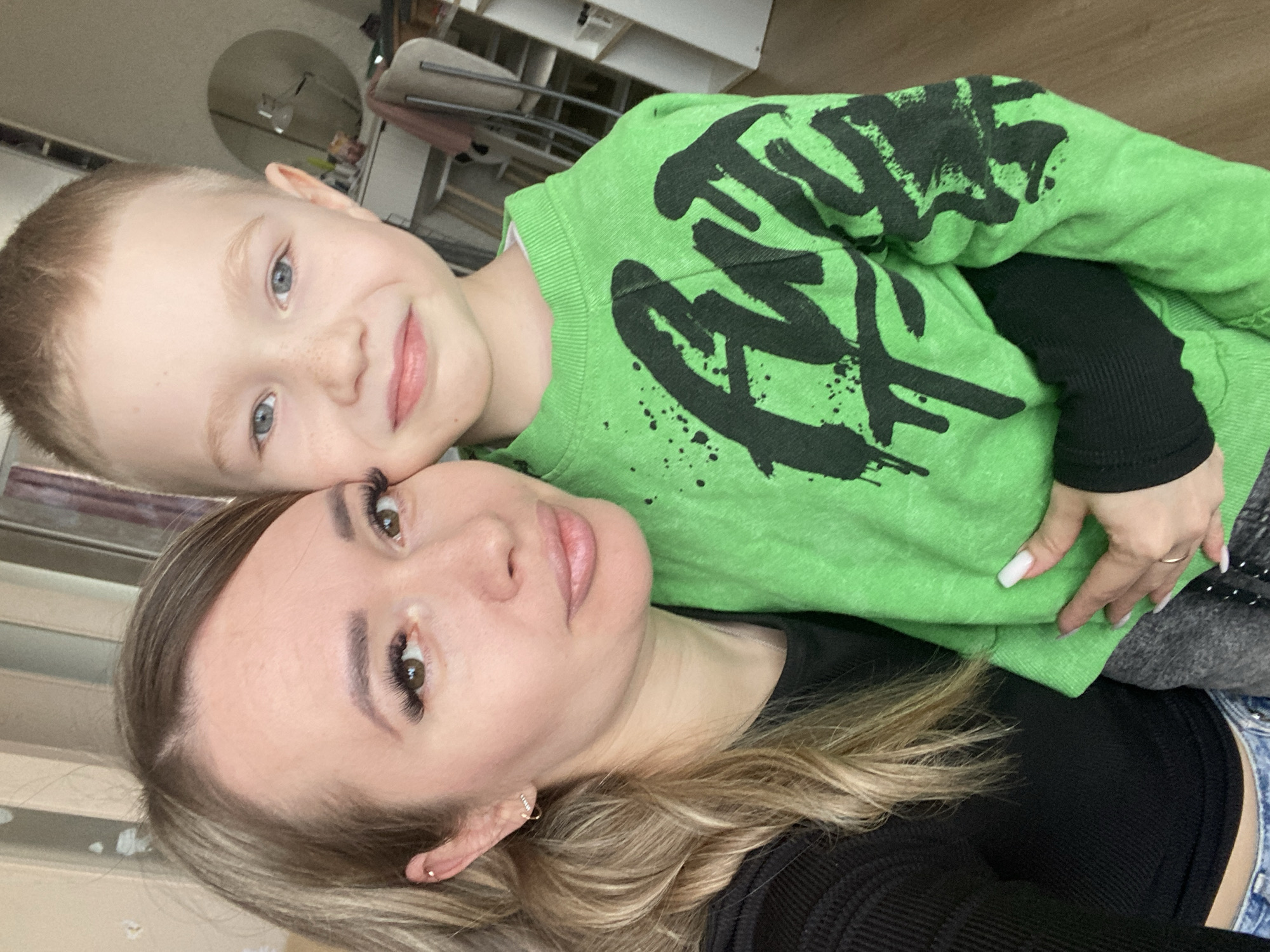 A woman and a young boy are in a selfie. The boy, with fair skin, blue eyes, and short blonde hair, is wearing a lime green sweater with a black graphic design. His face is positioned near the woman's. The woman has fair skin, brown eyes, and long blonde hair. She is wearing a black top. The background includes a room setting with furniture visible. The location is not specified in the image and the city of Izhevsk, Russia cannot be confirmed.