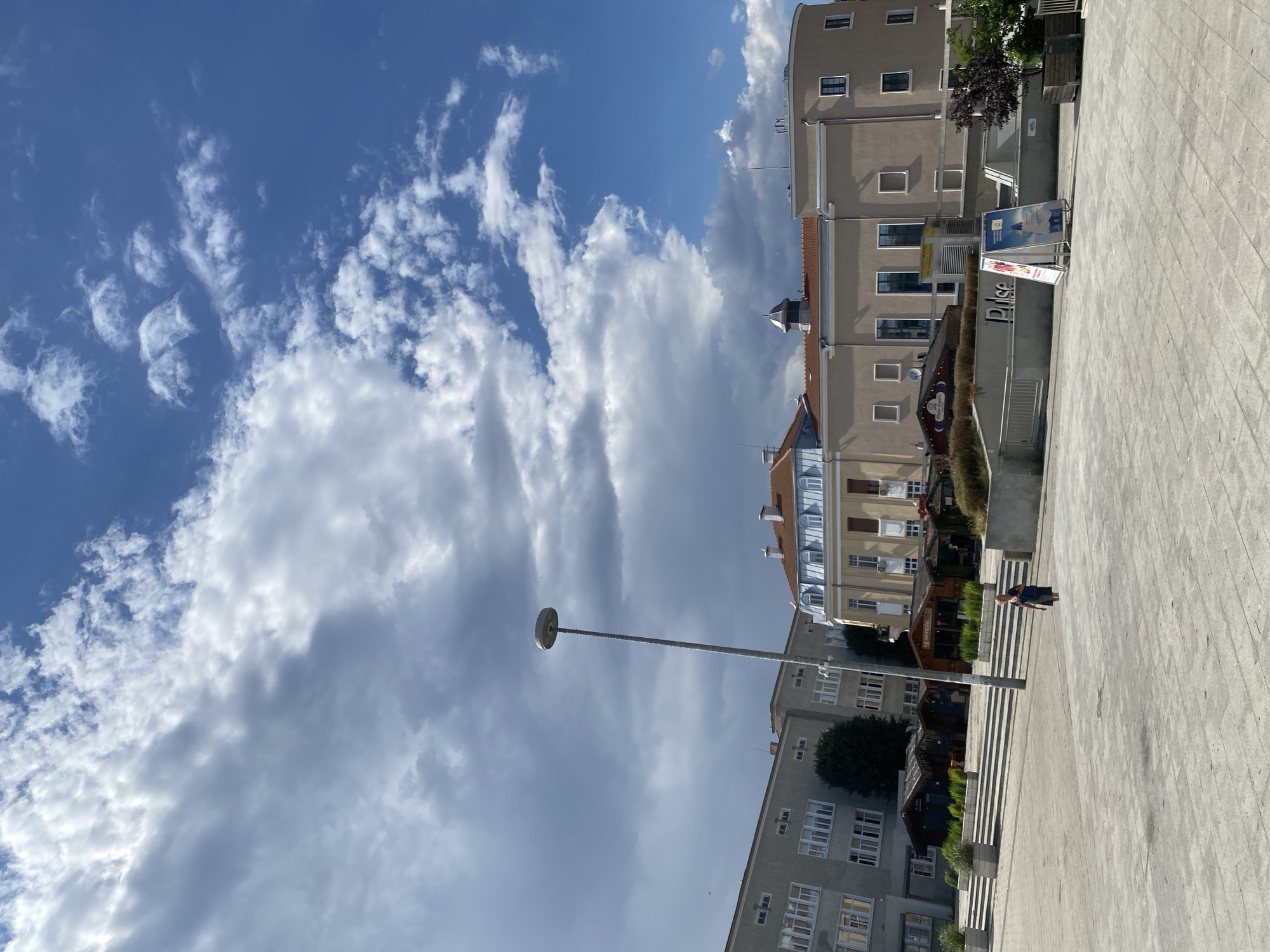 The image shows a wide, paved public square in Burgas, Bulgaria, under a partly cloudy sky. In the background, several buildings are visible. To the left, a multi-story, grey building with numerous windows dominates the scene. In the center, a tan building with a red-tiled roof and arched windows is prominent. Adjacent to this, on the right, is a rounded tan building with smaller, rectangular windows. Several small, dark-colored market stalls or kiosks are set up in front of the buildings, some with awnings. A tall, slender lamppost stands in the middle of the square. In the foreground, the square is paved with large, light-colored tiles. A lone woman wearing a dark dress walks across the square. In the mid-ground, a planter with green foliage is visible. Two A-frame signs are placed near the rounded building, one featuring an image of a lighthouse. The overall impression is an urban setting on a bright day with dramatic cloud formations.