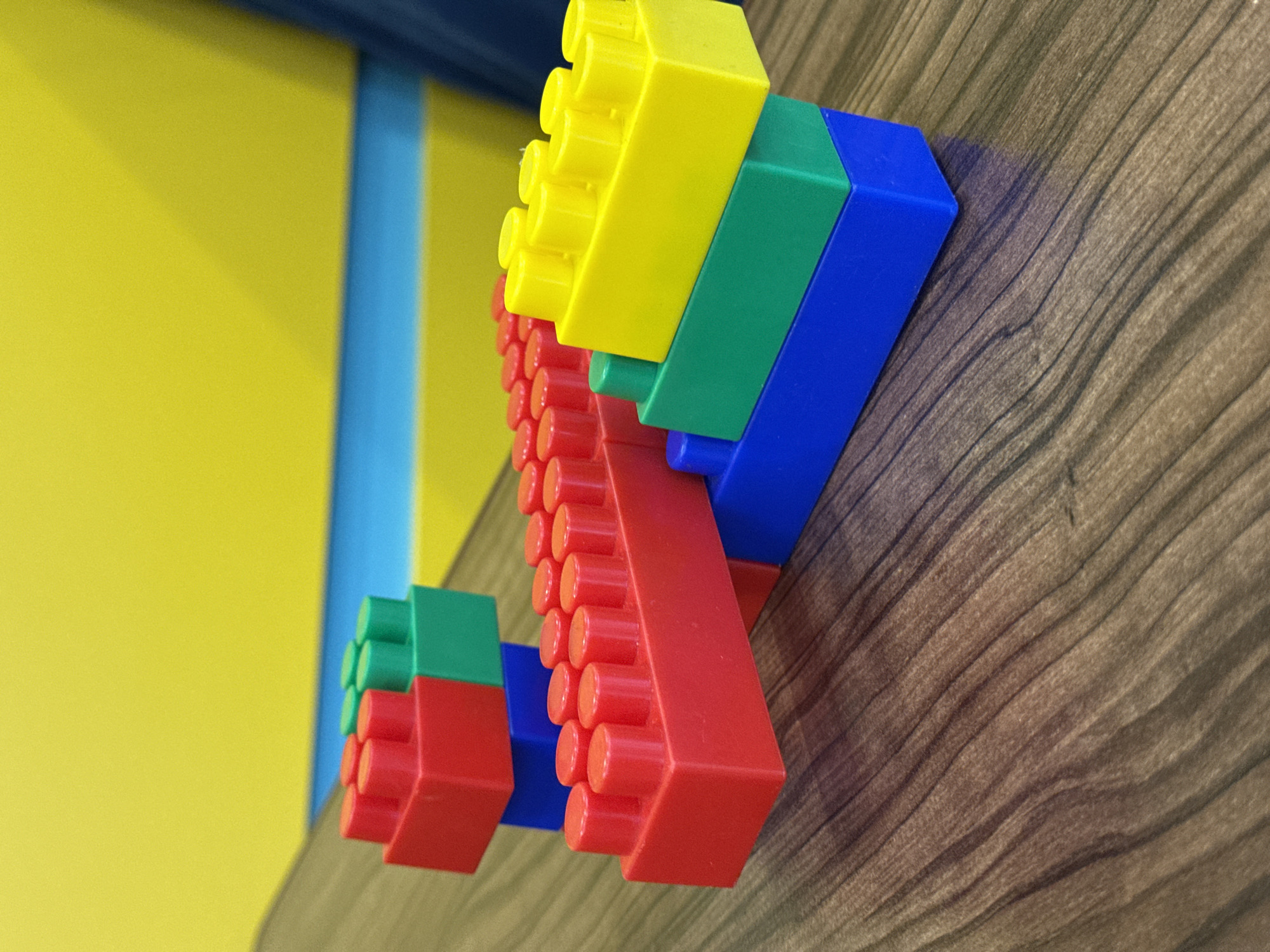 This image captures a vibrant display of large, colorful interlocking building blocks mounted on a wooden panel, set within an indoor environment in Perm, Russia. The blocks, reminiscent of children's construction toys, are primarily red, green, yellow, and blue.
Two distinct arrangements of these blocks are visible. The more prominent structure on the right consists of multiple red blocks at its base, topped by green blocks, then yellow blocks, and finally a blue block. This forms an ascending, stepped pattern, with the circular studs characteristic of such blocks clearly visible on their surfaces. A smaller, similar stack of red, green, and blue blocks is partially visible further to the left in the background.
The blocks are affixed to a dark brown wooden surface, which exhibits a noticeable wavy grain pattern. In the background, a bright yellow wall spans the left side of the frame, featuring a distinct vertical light blue stripe. A darker blue element is also present at the very top of the background. The lighting suggests an indoor setting, creating subtle shadows from the blocks onto the wooden surface. There is no visible text in the image. The scene conveys a playful and decorative interior.
