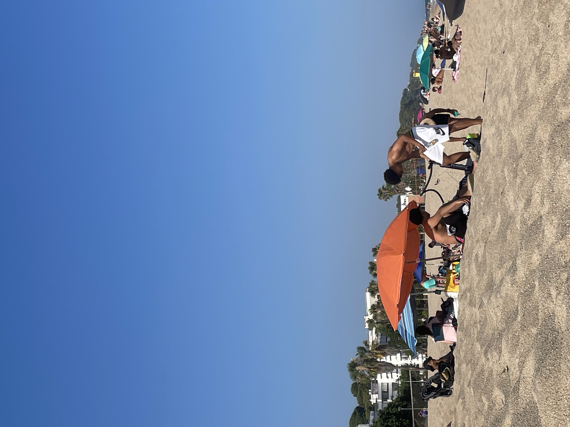 On a sandy beach in Canet de Mar, Spain, under a clear blue sky, several individuals are relaxing. In the foreground, two young men are standing on the sand. One, wearing white shorts with a black logo, is bent over examining something on the ground, while the other, partially visible, is seated. Behind them, several beach umbrellas provide shade for groups of people. An orange umbrella is prominent in the mid-ground, with a blue umbrella nearby. Further in the background, more umbrellas and beachgoers are visible, suggesting a populated beach. The coastline extends into the distance, with buildings and palm trees lining the shore. The sand is light brown and appears dry.
