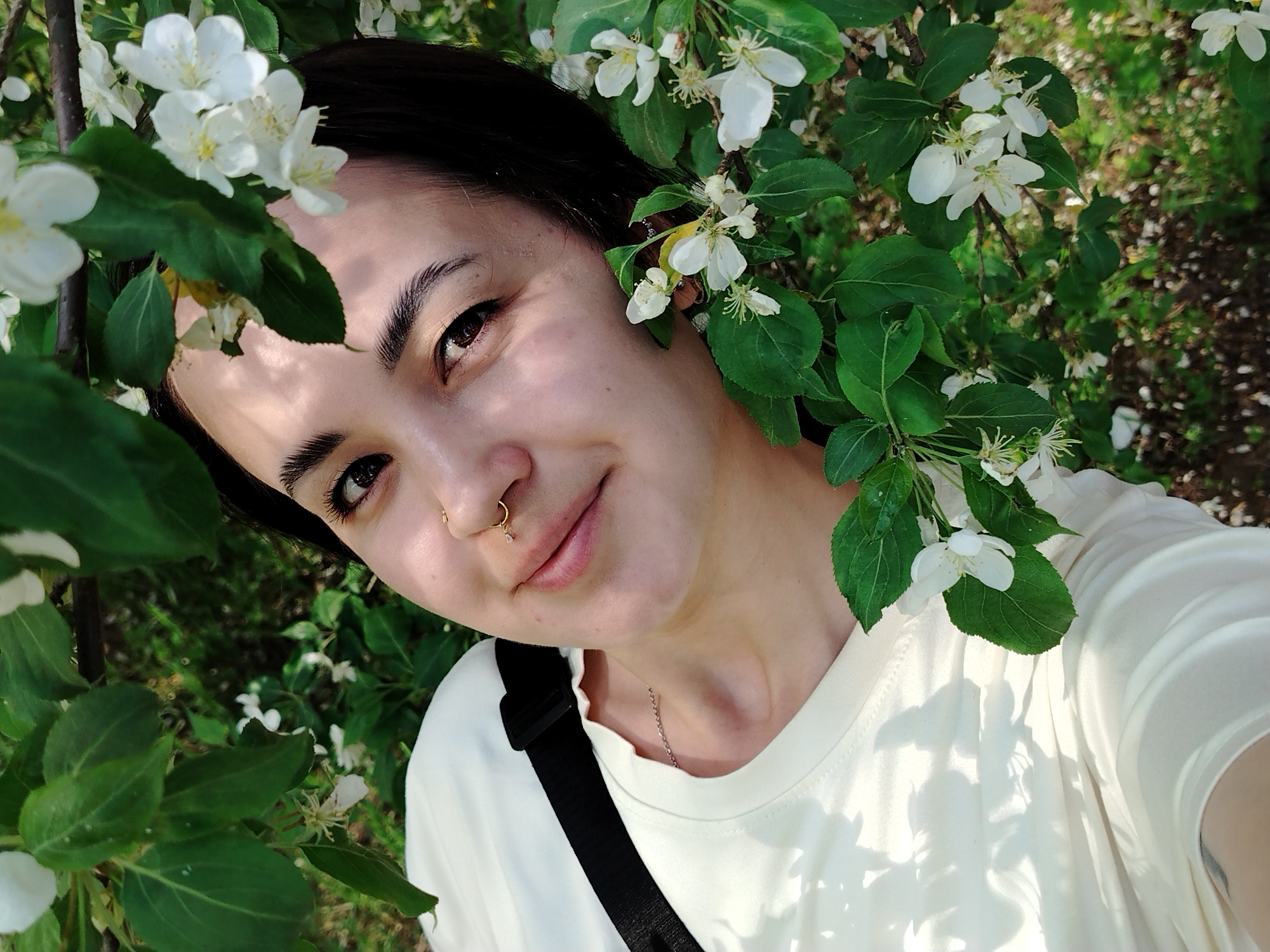 The image is a selfie of a young woman with dark hair, brown eyes, and a gold nose ring. She is surrounded by flowering branches with white blossoms and green leaves. She wears a light-colored T-shirt and a black strap crosses her body. The background is blurred, suggesting an outdoor location. The metadata indicates the location as Noviy, Russia.