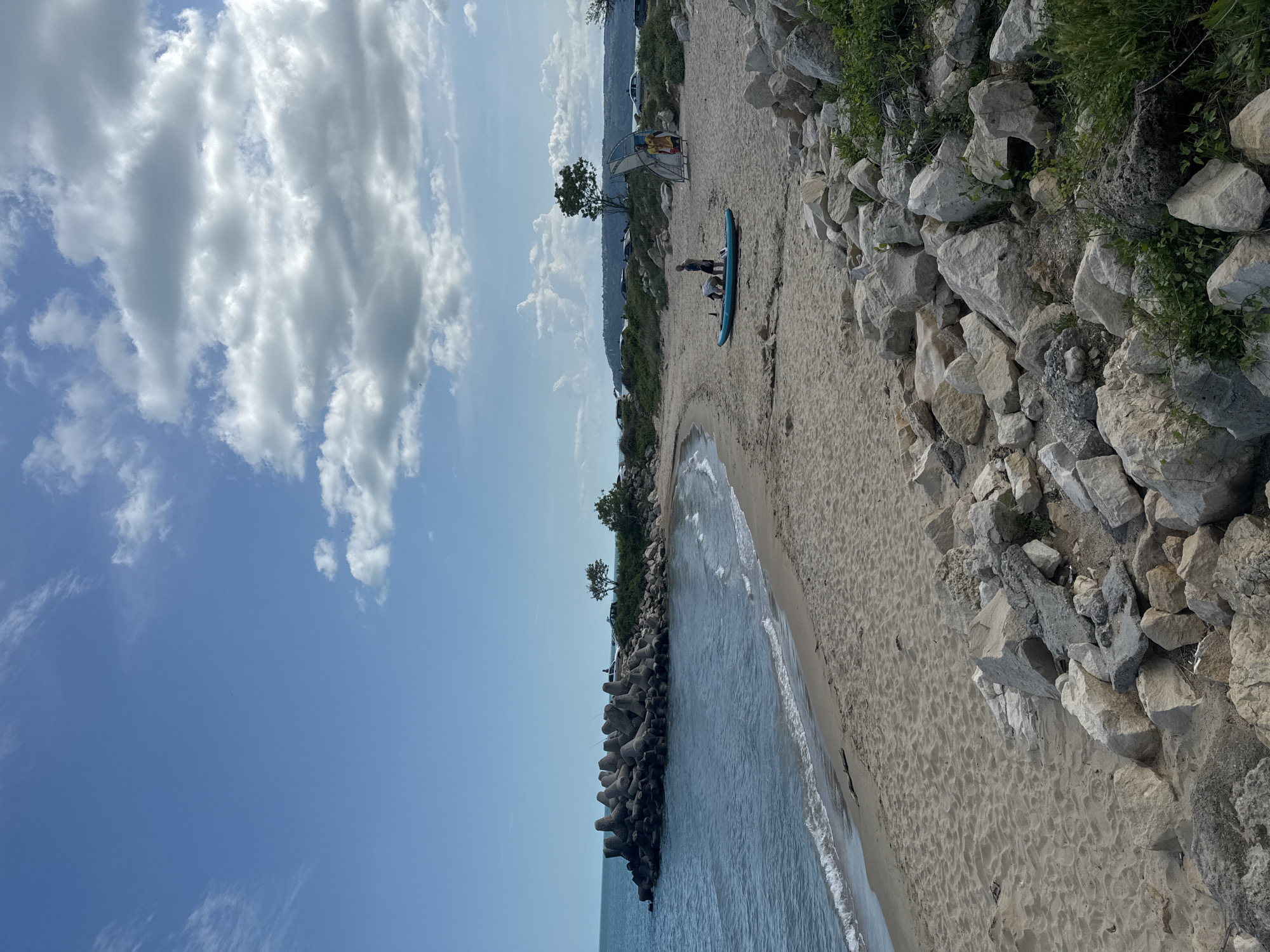 The photograph depicts a sandy beach in Varna, Bulgaria, with the sea on the left and a rocky outcrop on the right. A blue paddleboard lies on the sand, near two individuals, seemingly a man and a child. A partially visible wind shelter is behind them. In the background, further along the coastline, there are trees and parked vehicles. The sky is blue with scattered white clouds. The coastline is protected by concrete breakwater structures. The photo is taken at an angle, placing the horizon near the top of the frame.