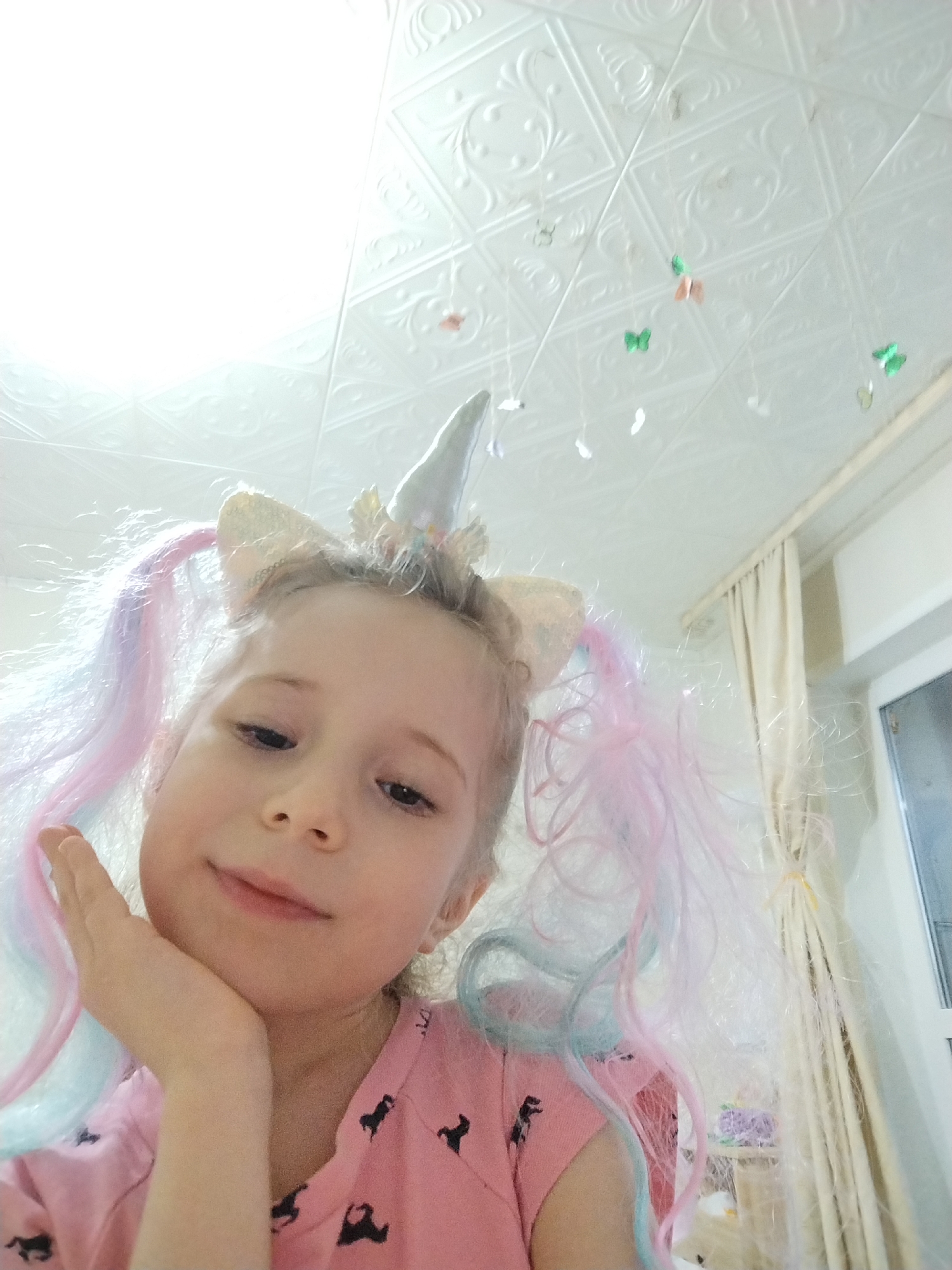 A child wearing a unicorn headband and a pink shirt with black horses is resting her chin on her hand. The location appears to be indoors, possibly a bedroom, in Марковский, Russia. The background includes a white textured ceiling decorated with hanging butterfly ornaments and a window with cream-colored curtains.