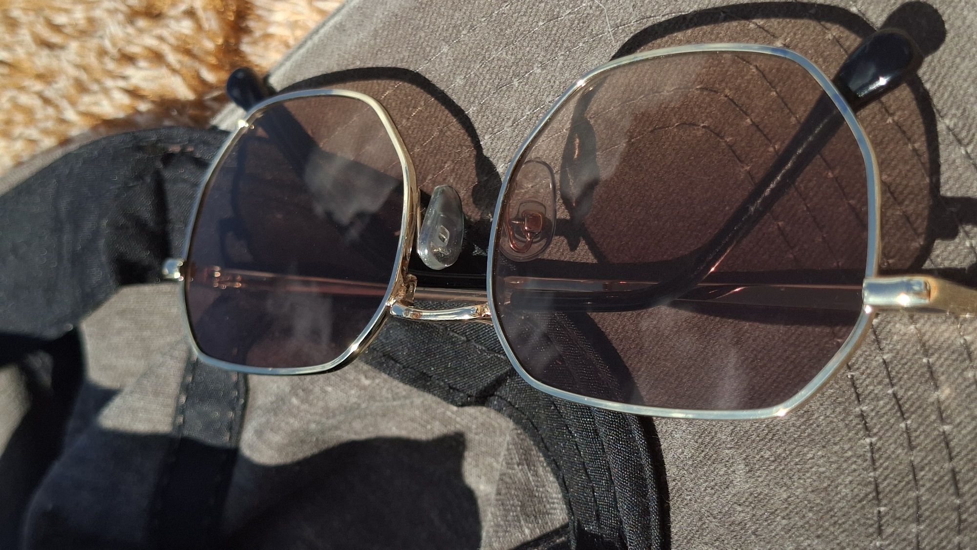 A pair of gold-rimmed sunglasses with brown lenses lies on a dark fabric surface, possibly a hat or clothing. The sunglasses are angled, with one lens showing more clearly than the other. The fabric is textured and appears to be outdoors, with sunlight creating shadows. The background is blurry and appears to be a rough, textured surface with a light brown color. The location is identified as Smalyavichy, Belarus.