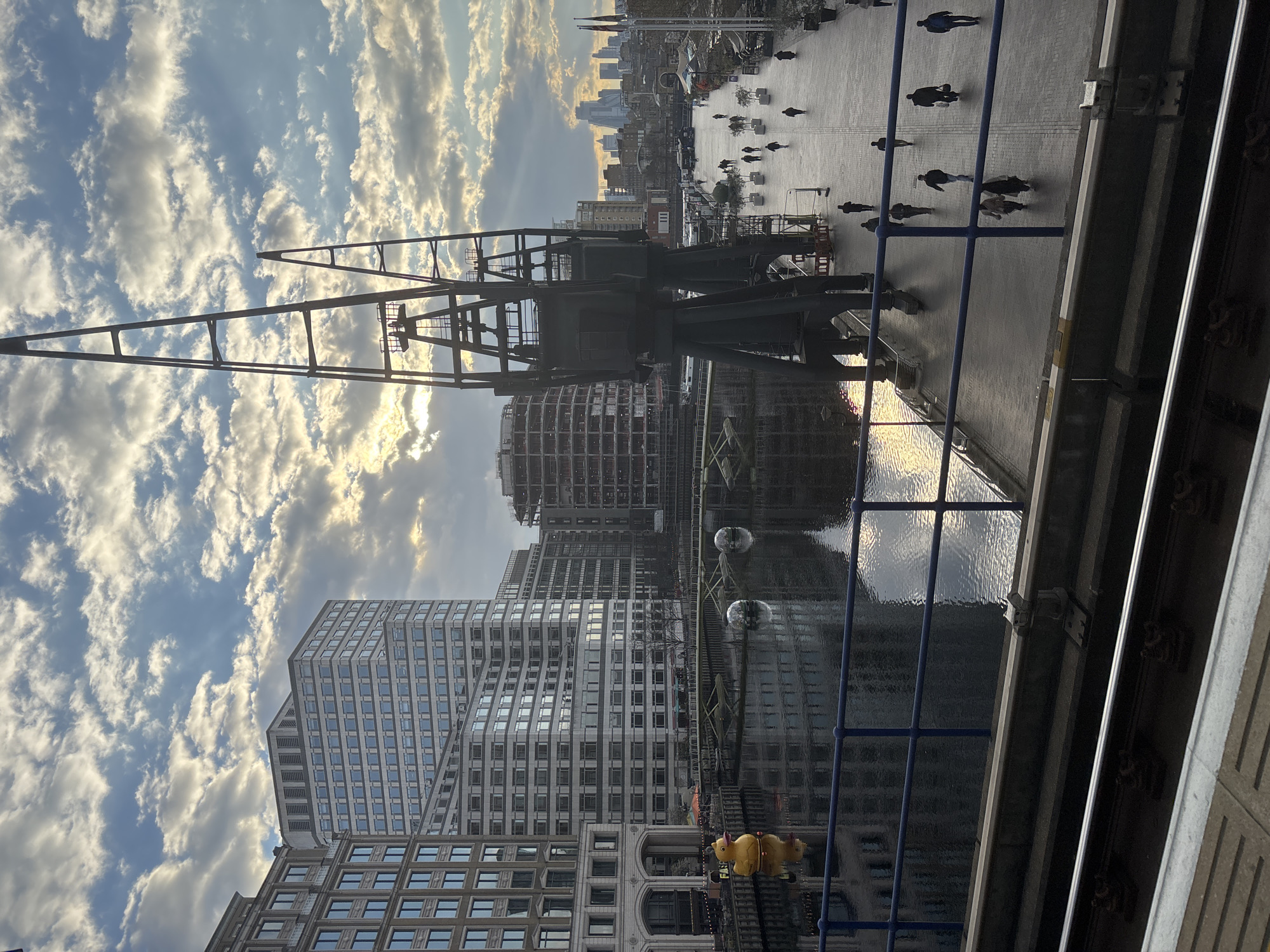 The image captures a view in the city of London, United Kingdom, likely near the docks or waterfront. A large industrial crane dominates the scene, its dark metal structure contrasting against the backdrop of a cloudy sky. Multiple modern buildings of various heights stand in the background, reflecting the city's contemporary architecture. A waterfront area is visible with people walking along a paved path. The water reflects the buildings and sky. A blue railing runs along the bottom right of the frame. There appears to be a pedestrian area with people strolling.