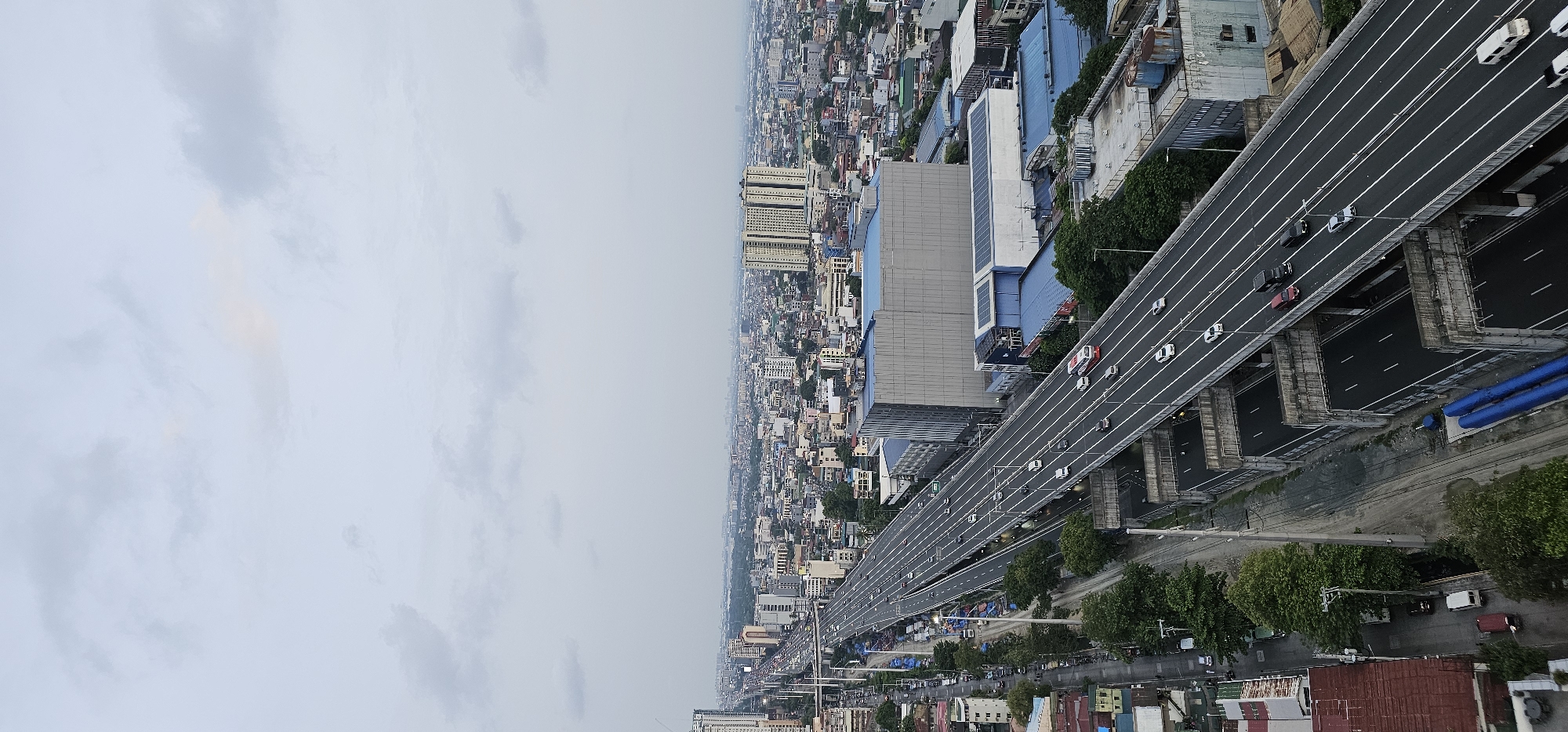 This is a vertical, high-angle, daytime aerial shot of a busy highway in Makati, Philippines. The highway is elevated and has multiple lanes of traffic, with cars and buses moving in both directions. The sky is overcast with light gray clouds. The highway is bordered by buildings and trees. In the foreground, there are some residential buildings with red roofs, and a street with a few vehicles. In the background, the cityscape stretches to the horizon, with numerous buildings of varying heights, including some tall skyscrapers. The overall scene depicts a dense urban environment with significant infrastructure for transportation.