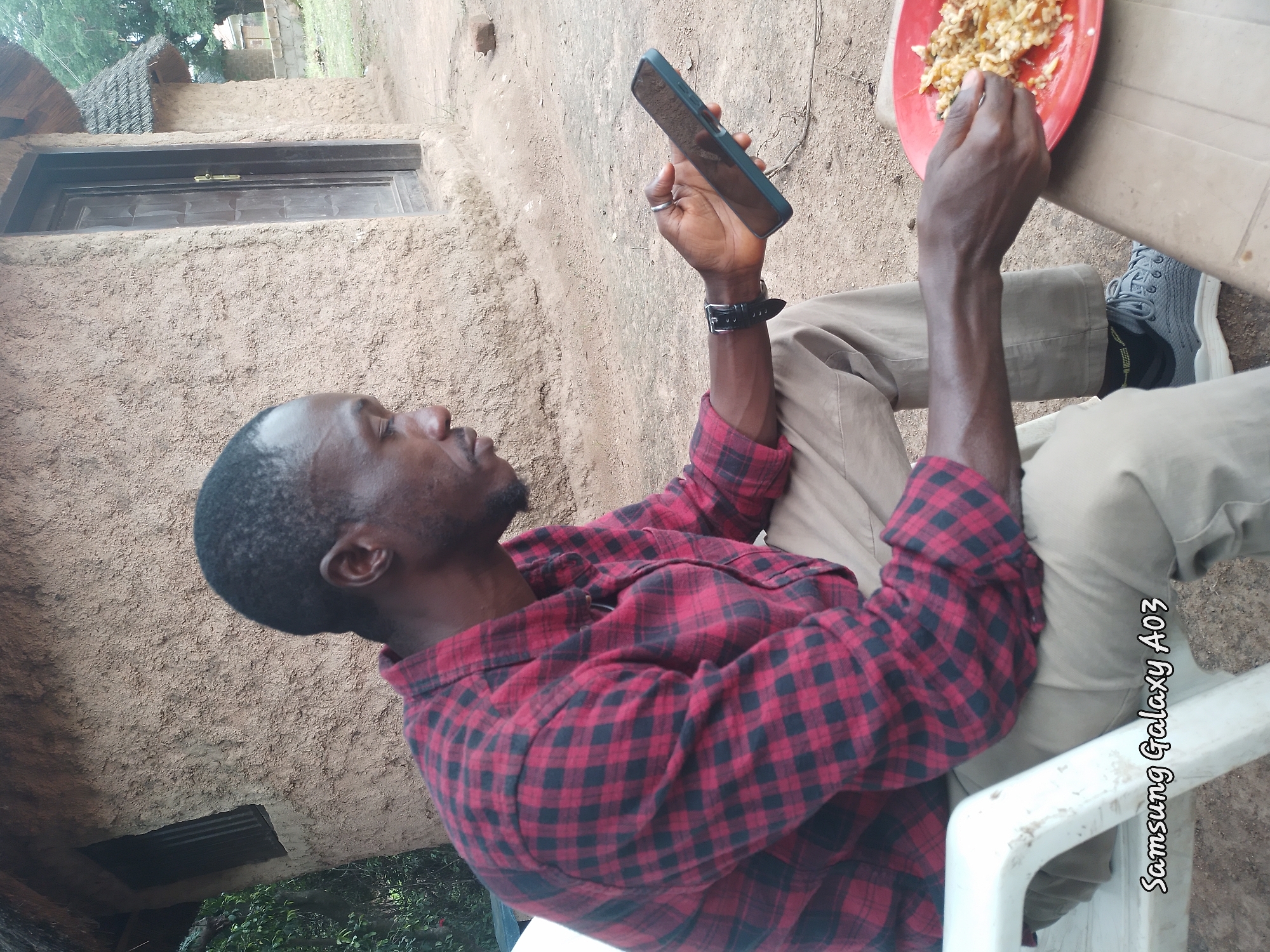 A medium shot captures a Black man seated in a white plastic chair outdoors, facing right and looking down at his smartphone. He is wearing a red and black checkered long-sleeved shirt and tan trousers. His left hand is raised, holding the phone, while his right hand is resting on a red plate containing rice. To his right, a beige plastic table holds the plate. In the background, a rustic building with a thatched roof and rough, brown walls is visible. The ground is dirt. The image is watermarked with "Samsung Galaxy A03" in white lettering at the bottom left. The scene appears to be a casual mealtime, possibly in a rural or traditional setting within or on the outskirts of Jos, Nigeria.