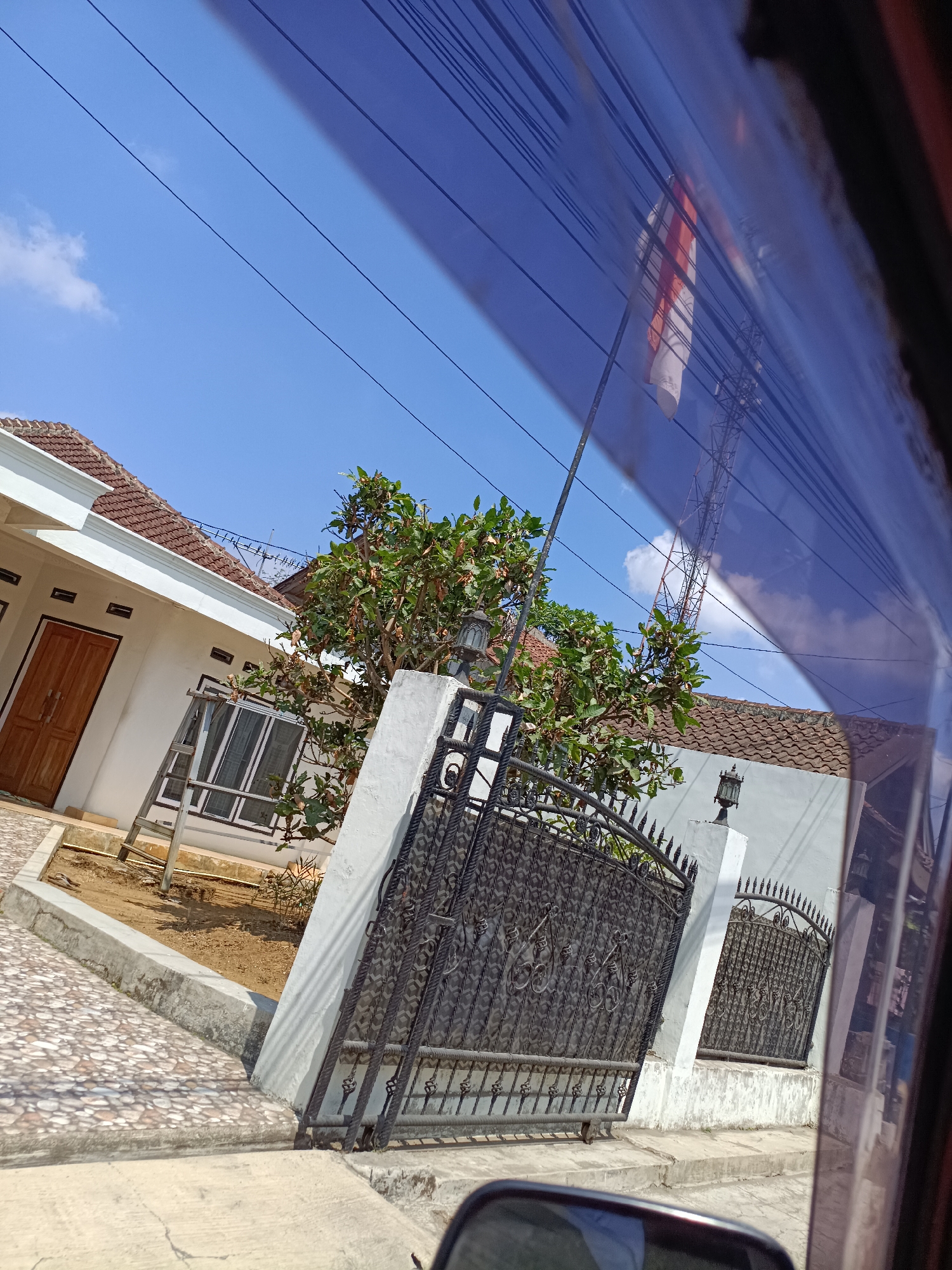 The image depicts a view from inside a vehicle, looking out through a tinted window. The scene captures a residential area in Bojong Genteng, Indonesia, under a clear blue sky with sparse white clouds. A white house with a tiled roof is visible on the left, featuring a wooden door and windows. In front of the house is a pathway paved with stones. Adjacent to the house is a decorative black metal gate, partially obscured by a tree with green foliage. A metal antenna tower is visible in the background, along with overhead power lines and a flag that appears to be red and white, possibly the Indonesian flag, on a pole. The reflection on the window shows additional power lines and a blurred image of the interior of the vehicle, including what looks like a rearview mirror. The environment appears to be a sunny day in a suburban or semi-rural setting.