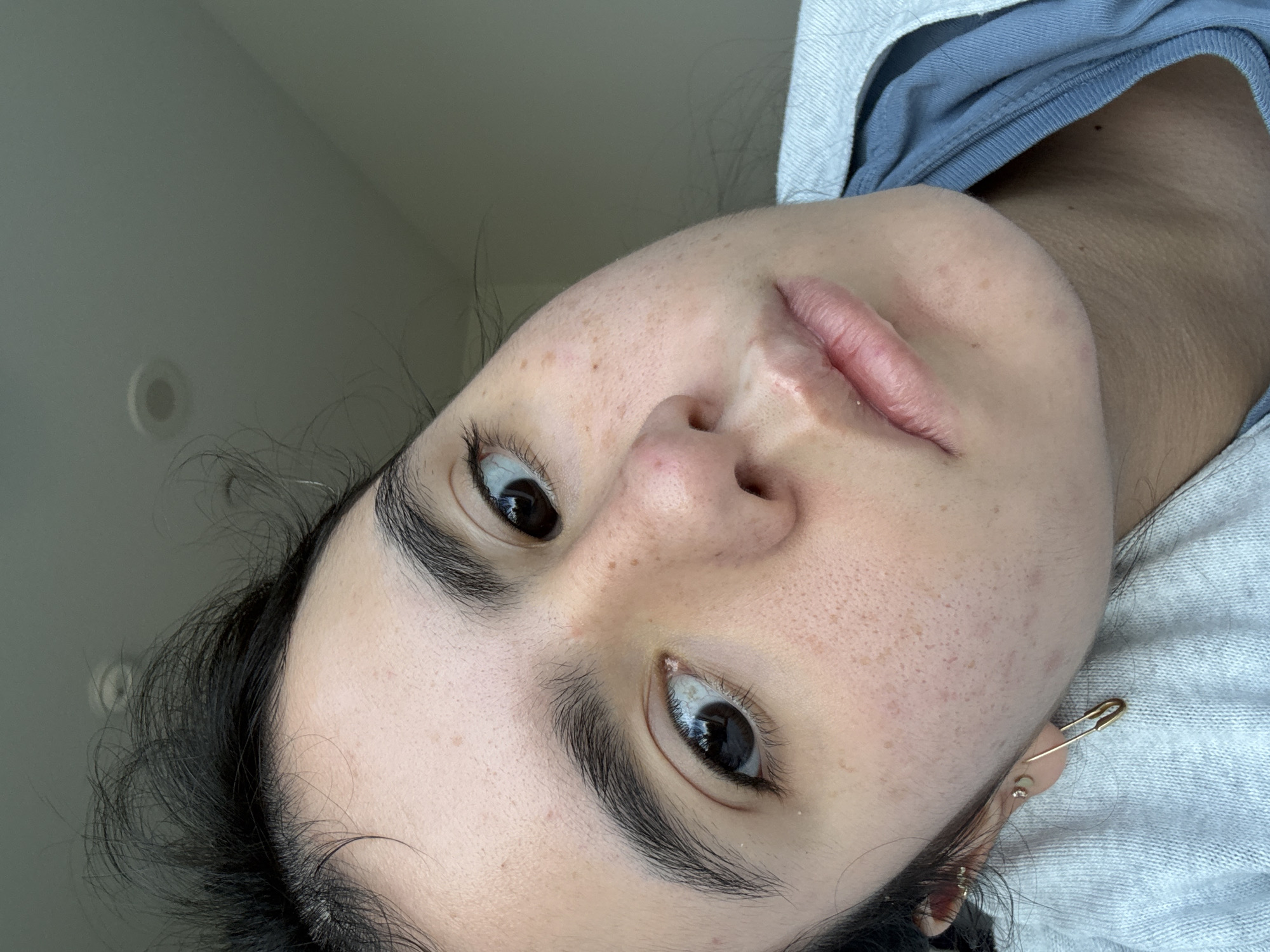 A close-up, angled portrait captures the face of a person, looking directly towards the camera. The image is taken from a low angle, showing the person's head tilted. They have fair skin with numerous freckles and small blemishes scattered across their nose, cheeks, and forehead. Their eyes are dark and appear to be looking slightly upwards, with visible dark eyelashes. Thick, dark eyebrows frame their eyes.
The person's dark hair is visible around their face, particularly near the forehead and temples, with some strands appearing slightly disheveled. Their lips are natural, with no visible makeup, and have a slightly chapped texture.
Clothing worn includes a light grey hooded garment, possibly a hoodie, with a blue t-shirt or top visible underneath at the neckline. A distinctive gold-colored safety pin earring is worn in the earlobe on the right side of the frame (the person's left ear), along with a few smaller stud earrings further up the ear. The background is a plain, light-colored surface, suggesting an indoor setting with soft, ambient lighting. There is no visible text in the image.