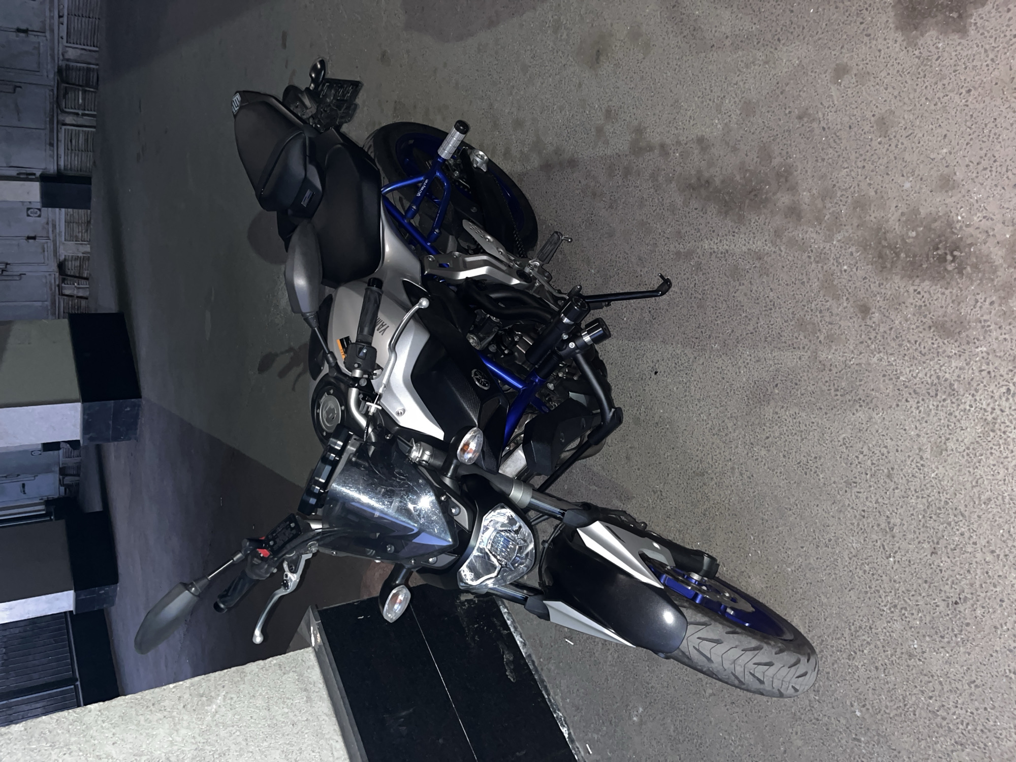 The image shows a Yamaha MT-07 motorcycle parked on a grey concrete surface outdoors. The motorcycle is primarily silver and black with blue accents on the frame and wheels. It has aftermarket crash bars installed. The location appears to be an urban environment, possibly a parking area or a street adjacent to a building with what appear to be storage units or metal lockers visible in the background. The lighting suggests it is night or a dimly lit area. Based on the image, there is no concrete evidence to confirm the location is specifically in Saint Petersburg, Russia.