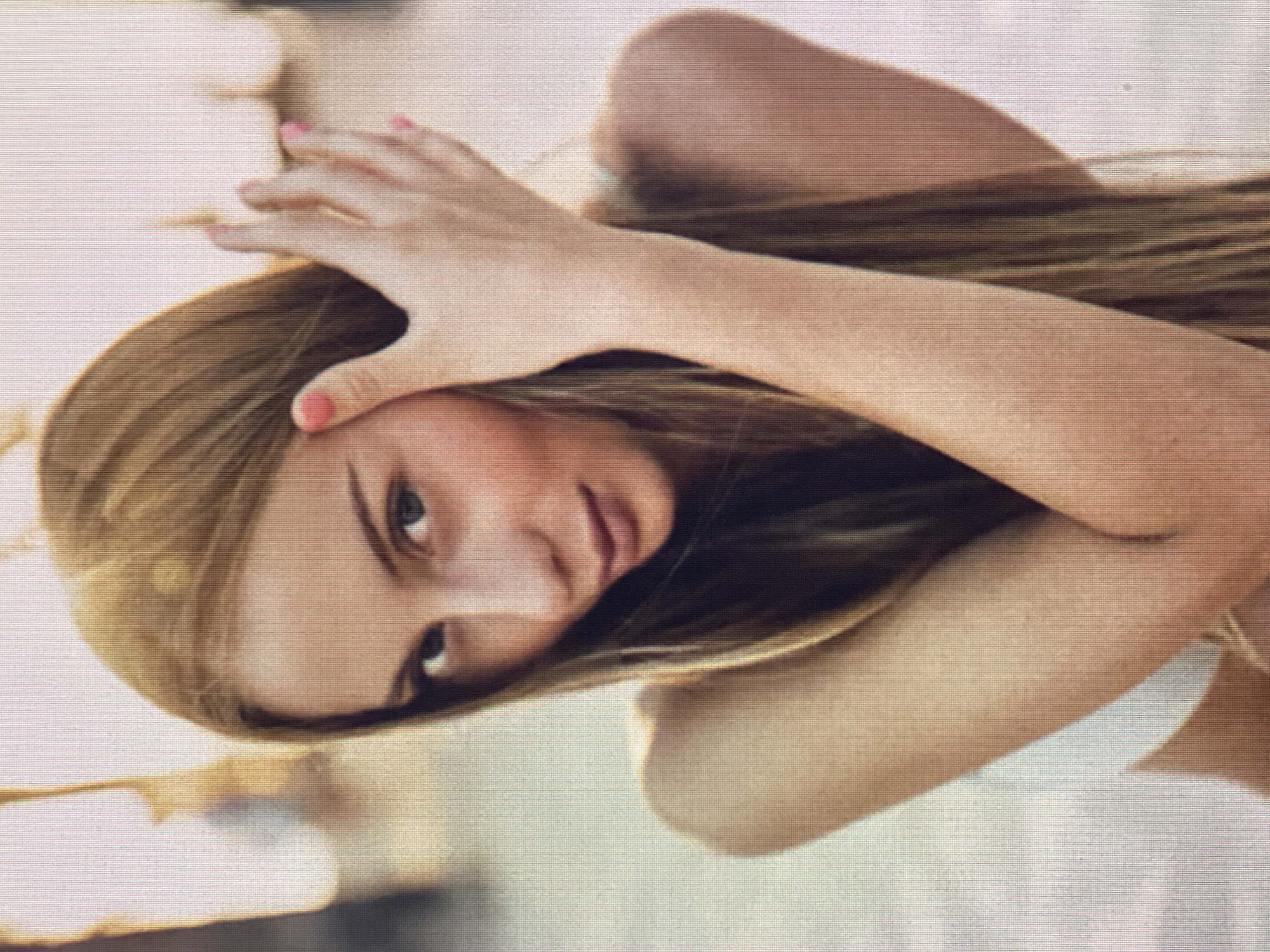 A young woman with long blonde hair is pictured in a close-up, outdoor shot, with a blurry, bright background that suggests a sunny day in a city setting. She is looking directly at the camera, her right hand resting on the left side of her head, with her fingers splayed. Her nails are painted pink. She is wearing a white garment. The image is pixelated, giving it a retro or vintage aesthetic. The lighting suggests that the sun is behind her, creating a halo effect around her hair.