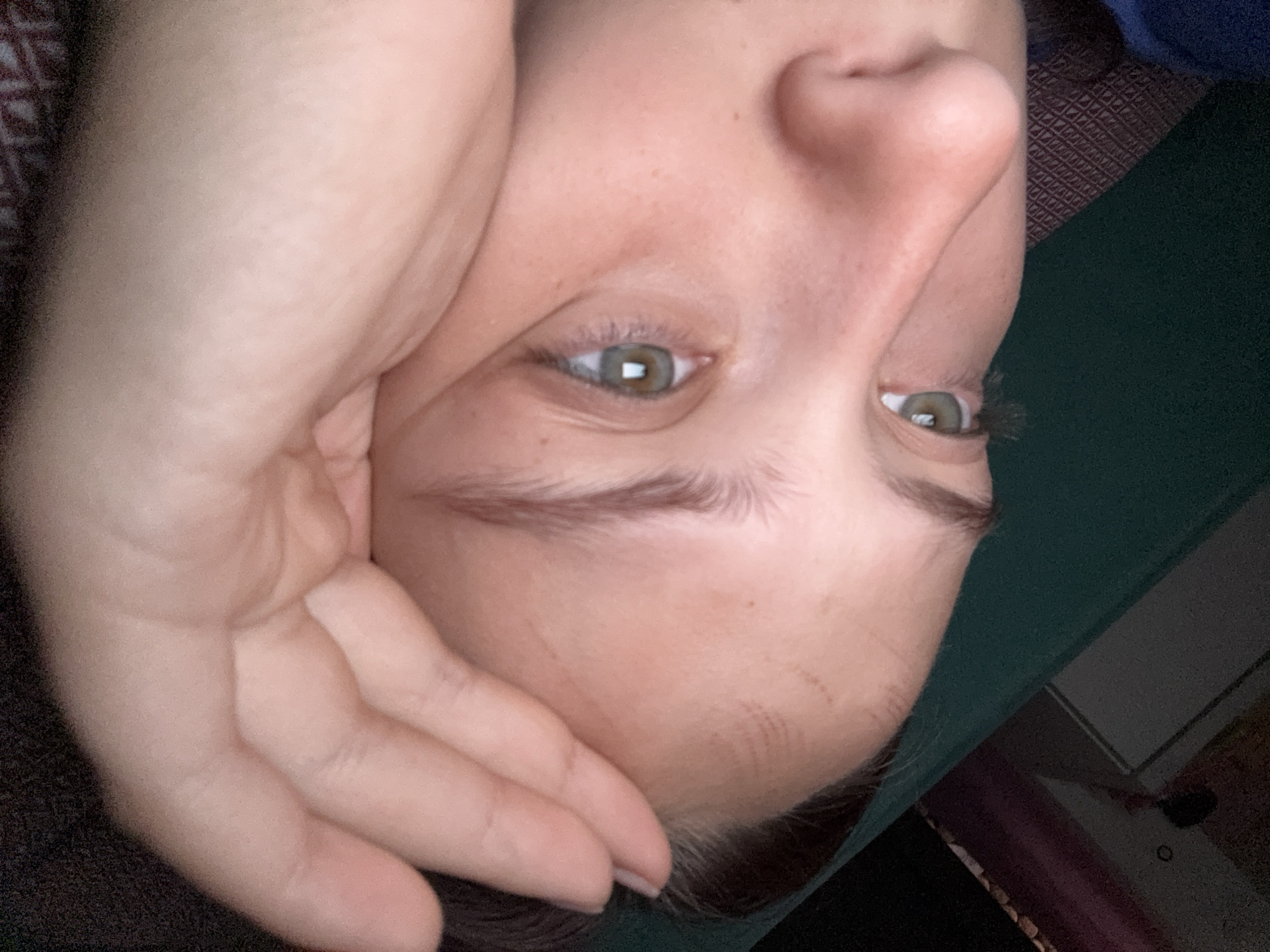 A close-up, slightly rotated portrait shows the face of a person, seemingly lying down. The dominant features are their open hazel or green eyes, which show a rectangular reflection suggesting a screen. Their nose and part of their forehead are also visible. A hand rests against the side of their face and forehead. The skin has a fair complexion with visible pores and light freckles. Faint red marks, possibly impressions from fabric or resting, are present on the forehead. The person's eyebrows are well-defined.
The background is largely out of focus, indicating an indoor setting. Dark, possibly patterned fabric is visible on one side, while a dark green surface, perhaps a blanket or mat, occupies another part of the background. A very blurry glimpse of what appears to be a room interior, possibly a doorway or furniture, can also be discerned. The person appears to be in a state of rest or quiet contemplation. No text is visible in the image. The scene is set in Прикамский, Russia.