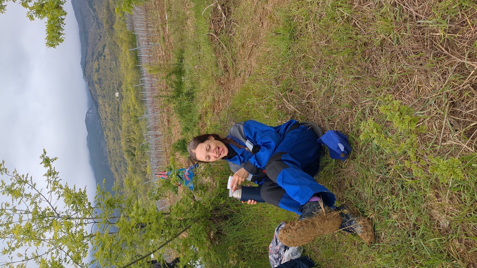 A woman in a blue jumpsuit is sitting on the ground in a grassy field, holding a thermos. She has brown hair in a braid and is sticking her tongue out. Her shoes are covered in mud. A tree is to her left, and a vineyard is behind her. Mountains are visible in the distance under a cloudy sky. The location is likely outdoors, potentially rural or agricultural, and not confirmed in Pushkino, Ukraine.