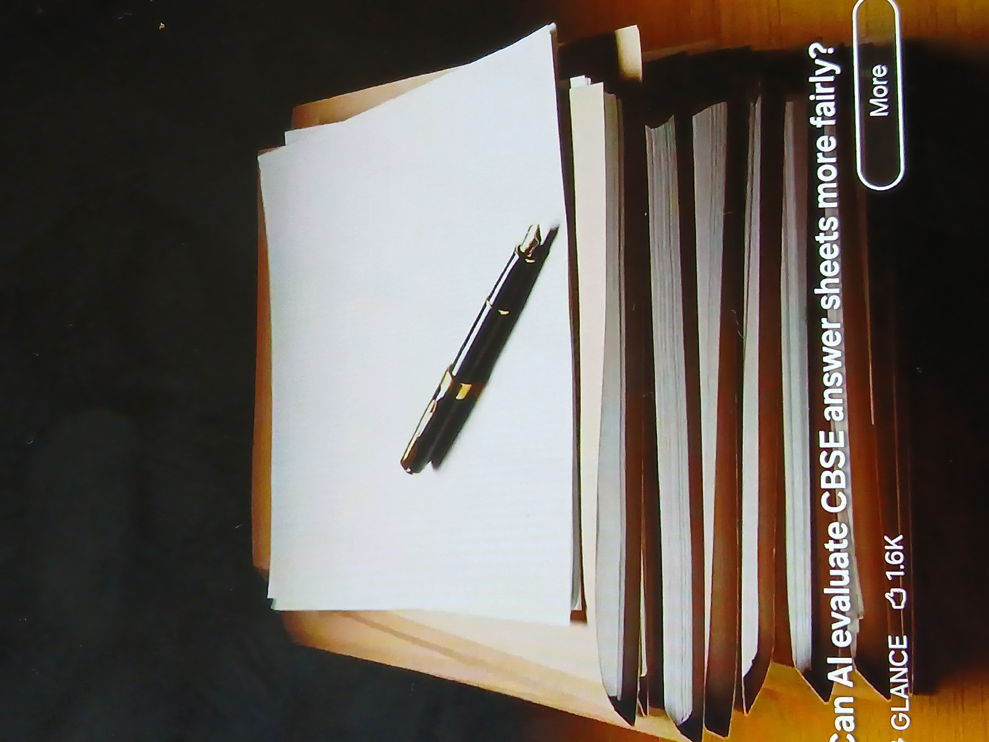 Here's a detailed description of the image:
The image shows a stack of papers and books on a wooden surface. A black fountain pen with gold accents lies on top of the papers. The papers are mostly white and appear to be blank. The books are stacked on the right side of the image, and their pages are visible.
The text on the bottom right of the image reads: "Can AI evaluate CBSE answer sheets more fairly? GLANCE 1.6k." There is a "More" button to the right of this text.
Given the text references "CBSE answer sheets," it's highly likely the image relates to education in India. However, there's no information in the image itself to confirm the location is Bengaluru or any other specific city.