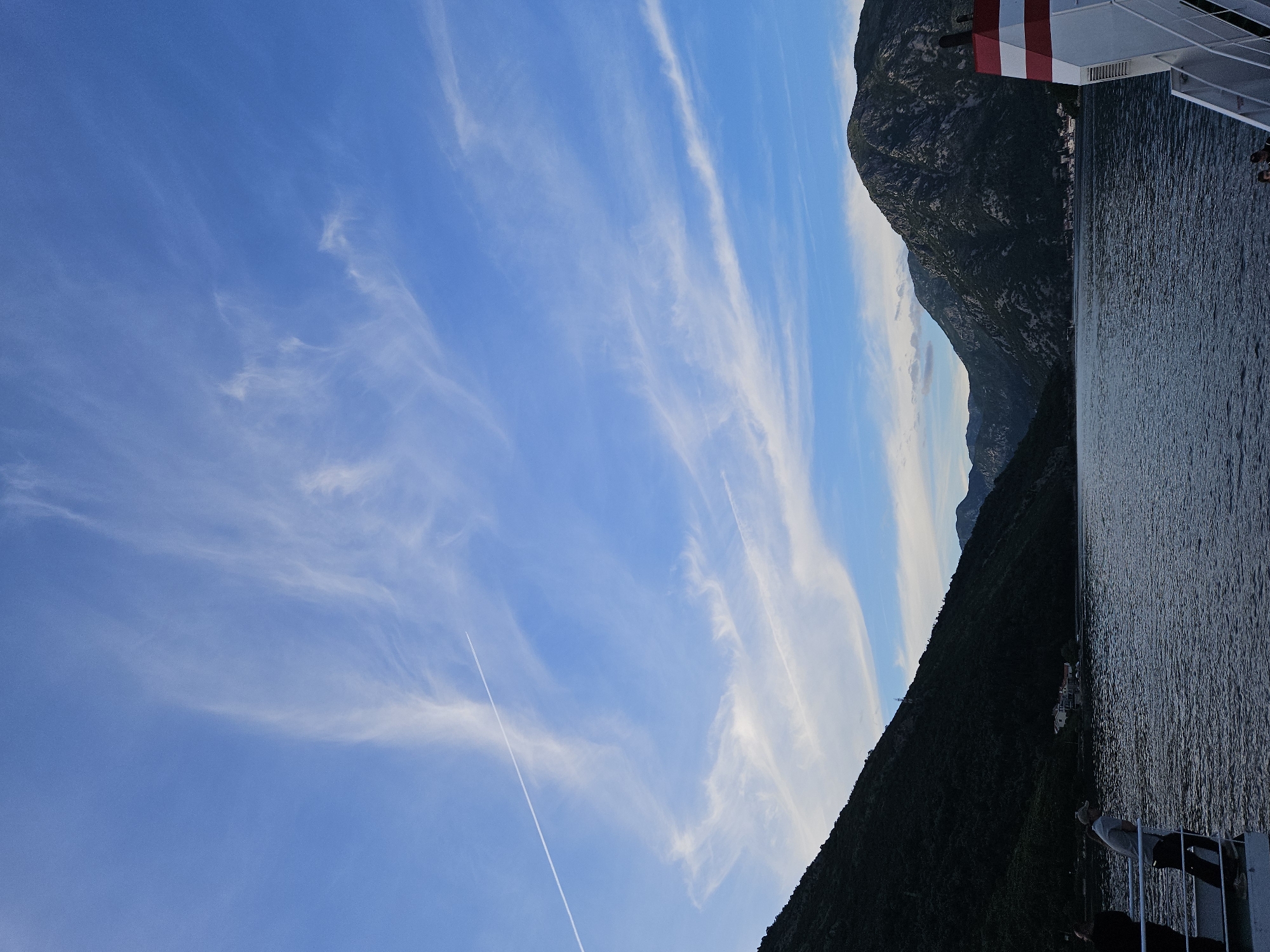 The image shows a landscape featuring mountains and water under a blue sky with wispy clouds and a contrail. A portion of a vessel, likely a ferry or ship, is visible in the top right corner. The scene is possibly taken from a boat moving through a fjord. The exact location is unknown.