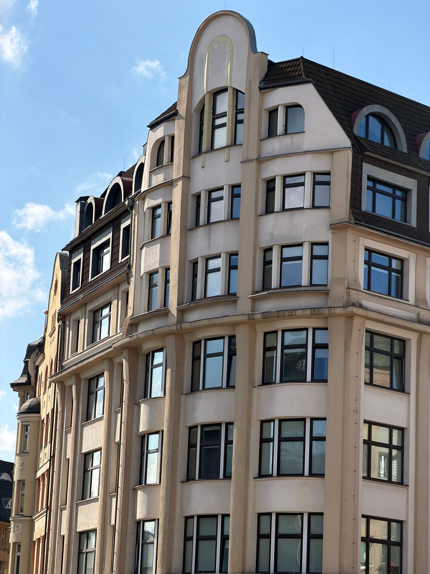 A close-up, eye-level, medium shot captures the upper floors of a cream-colored Art Nouveau building with a dark tiled roof, set against a clear blue sky with a few wispy white clouds. The building features multiple rows of windows with black frames, some rectangular and some arched. Decorative elements include curved cornices and vertical pilasters. To the left, portions of a slightly darker, narrower building with a peaked roof are visible, and further left, a sliver of a building with a small rounded tower is seen. The architectural style suggests an urban setting.
While the image itself does not contain explicit clues to its location, this building is located on Ulica Wita Stwosza in Wrocław, Poland. The specific address is Wita Stwosza 33.