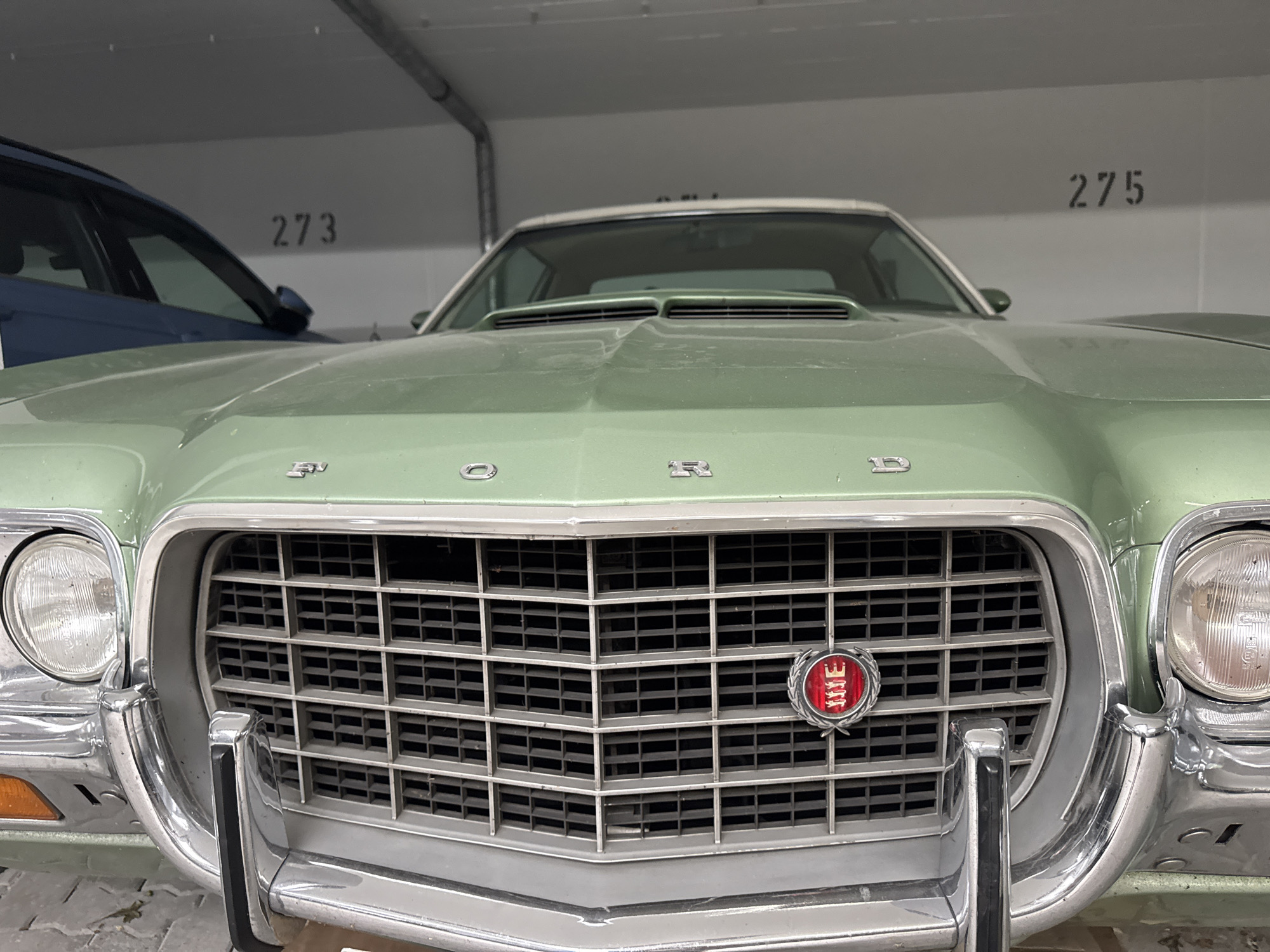 The image displays the front of a light green vintage Ford automobile, likely a Gran Torino based on the grille and emblem. The car is parked indoors, possibly in a multi-story car park or garage, as indicated by the ceiling structure and numbered parking spots visible on the wall in the background. The numbers "273" and "275" are clearly visible on the wall, suggesting a specific location within a numbered parking facility. The car features a prominent chrome grille with a red and silver emblem in the center, round headlights, and chrome bumpers. To the left of the Ford, a portion of a dark blue vehicle is visible. The overall lighting suggests an indoor environment with overhead illumination. The image was taken in Höhenkirchen-Siegertsbrunn, Germany.