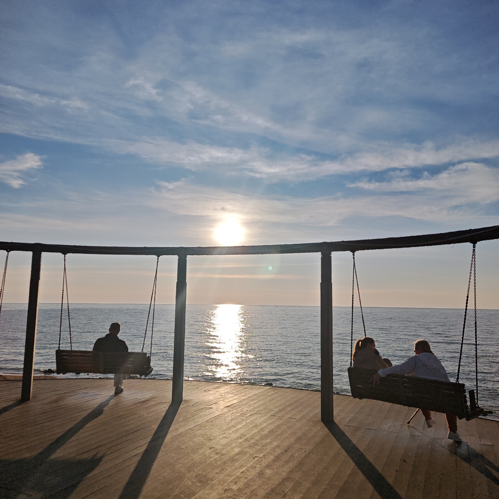 The image shows a seaside scene in городское поселение Щелкино, Ukraine. A circular swing set with three swings is positioned on a wooden platform overlooking the sea. The sun is setting or rising over the water, creating a bright reflection on the sea surface. A person sits alone on the swing on the left. Two people are seated on the swing on the right. The sky is blue with scattered white clouds. The scene is bathed in sunlight, casting shadows from the swing set posts onto the wooden platform. The sea is calm.