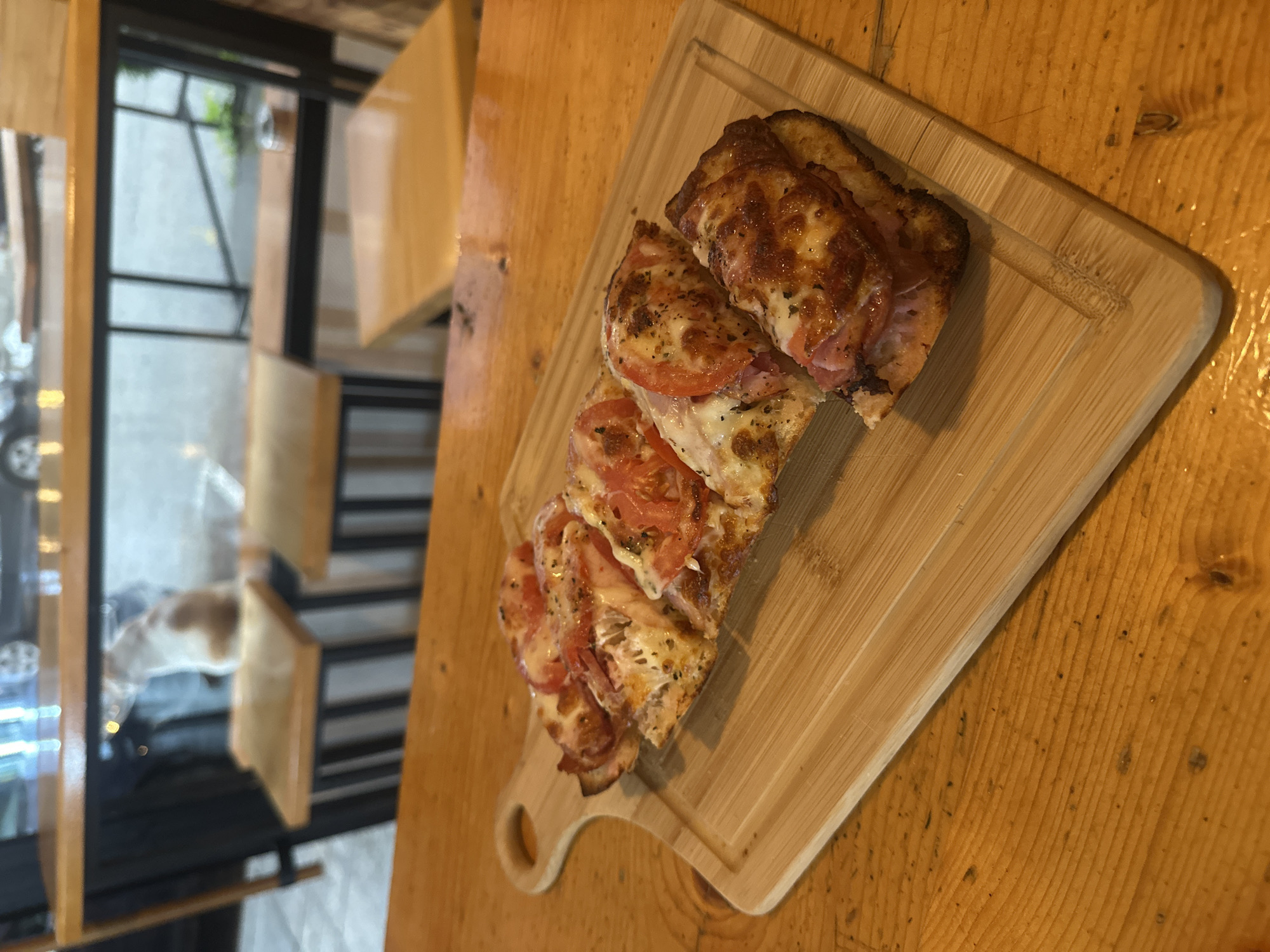 The image shows a partially eaten sandwich or flatbread on a light-colored wooden cutting board with a handle. The sandwich consists of several slices, topped with melted cheese, tomatoes, and possibly ham or another kind of meat. The cutting board is placed on a light brown wooden table. In the background, a window is visible with a blurry view of an outdoor urban scene. Based on the presence of the Cyrillic alphabet on a street sign visible through the window, the location is likely in Bulgaria. Given the architecture and information available, this is possibly Veliko Tarnovo, Bulgaria. However, without more specific details, the exact location within Veliko Tarnovo is uncertain. The focus of the image is on the sandwich, suggesting food photography.