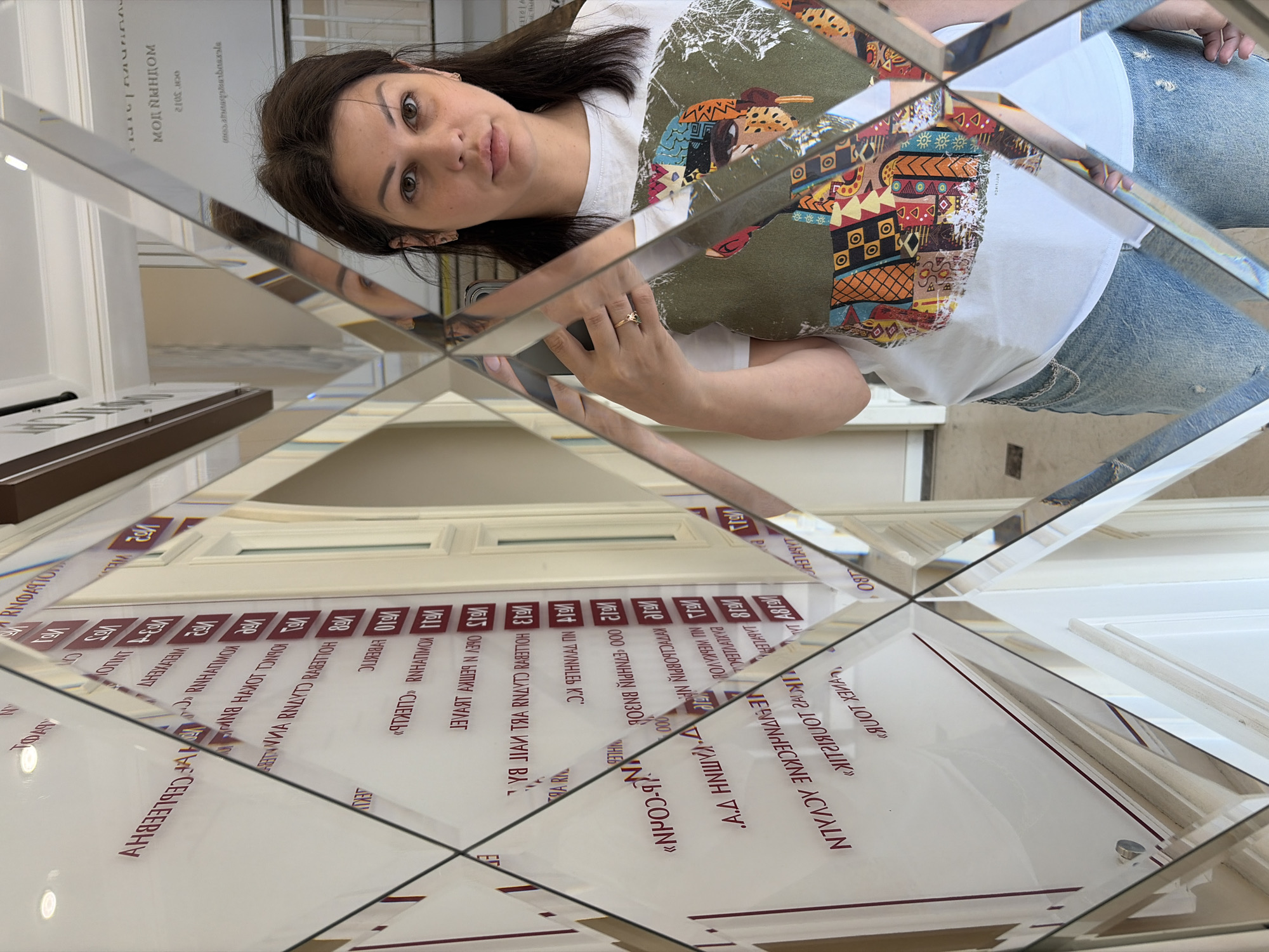 A woman is depicted taking a mirror selfie in an interior location, identified as Sochi, Russia. The woman, with dark hair and a neutral expression, wears a white t-shirt featuring a colorful graphic design of a stylized head or figure on an olive green background, paired with light blue distressed denim jeans and a waist chain. She holds a smartphone with a silver/grey case in her right hand, displaying a ring with a colorful stone on her right ring finger. The environment consists of a wall covered entirely in beveled, diamond-shaped mirror tiles, creating multiple fragmented and reversed reflections of the surroundings.
Reflected in the mirrors is a white wall with vertically aligned dark red text, appearing to be a directory or list. Visible text includes numbers "№1" through "№15", along with various Russian names and company names such as "МОЙ ЯНДОМ" (My Yandom/My Home), "2105.co", "СЕБЕЛЕВА Н.А.", "КОРНЕЛЕВА Р.П.Н.", "ЯНА ЛАЙФ", "ИНЖИНИРИНГ", "НПО-КОРН", "ТУРБУКС", "ГАЛАНТ", "ФАБЕРЛИК", "СПЕКТР", "ТРЕЙД", "МЕТАЛЛ", "А.А. ШИШКИНА", and "ТУРИСТИК". Architectural details reflected include white door frames or moldings, a white ceiling with recessed lighting, and a light-colored tiled floor. The mirror's facets distort and fragment the woman's image and the background text.
