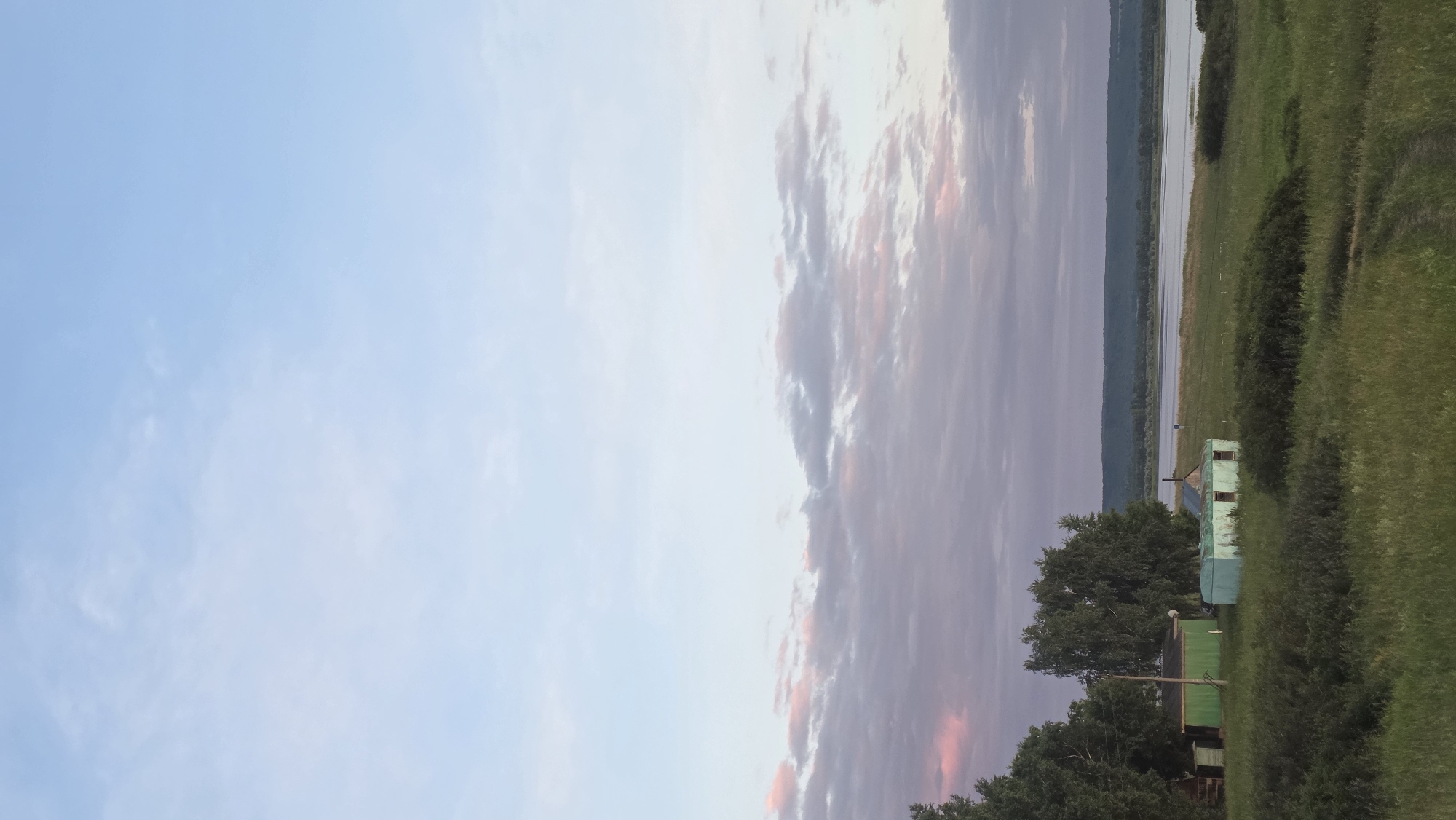 This is a vertical, eye-level shot of a rural landscape near the city of Alga, Russia. The upper two-thirds of the image are dominated by a pale blue sky with wispy, muted clouds that show hints of pink and purple, suggesting either sunrise or sunset. In the lower third, a grassy field stretches towards a wide, calm body of water that reflects the sky. Along the edge of the field, near the left side of the frame, are two small, weathered structures. The structure on the left is painted a dark green, and the one to its right is a faded turquoise color with visible windows. Dense green foliage, including trees and bushes, frames the left side and foreground of the scene. A faint dirt path is visible in the grass on the right side of the foreground. The horizon line is low, emphasizing the vastness of the sky and the expanse of the water. There are no people or animals visible in the image.