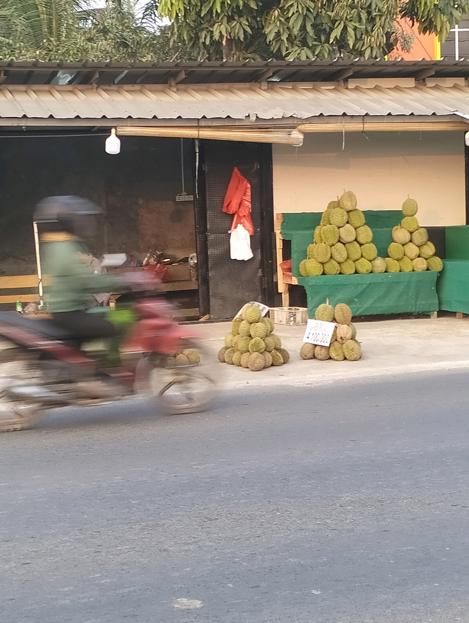 The image depicts a street scene in Perumnas Way Kandis, Indonesia. A person, wearing a green jacket and helmet, is blurred due to motion as they ride a red motorcycle from left to right across the frame. In the background, a stall or shop is visible with a corrugated metal roof. A single light bulb hangs from the ceiling of the stall. Red and white plastic bags are hanging on the right side of the stall. In front of the stall, there are two large stacks of durian fruit. A smaller stack of durian is also present in the foreground, to the left, with a white sign displaying handwritten text that appears to be a price. The ground is paved with asphalt. Trees are visible above the stall's roof.