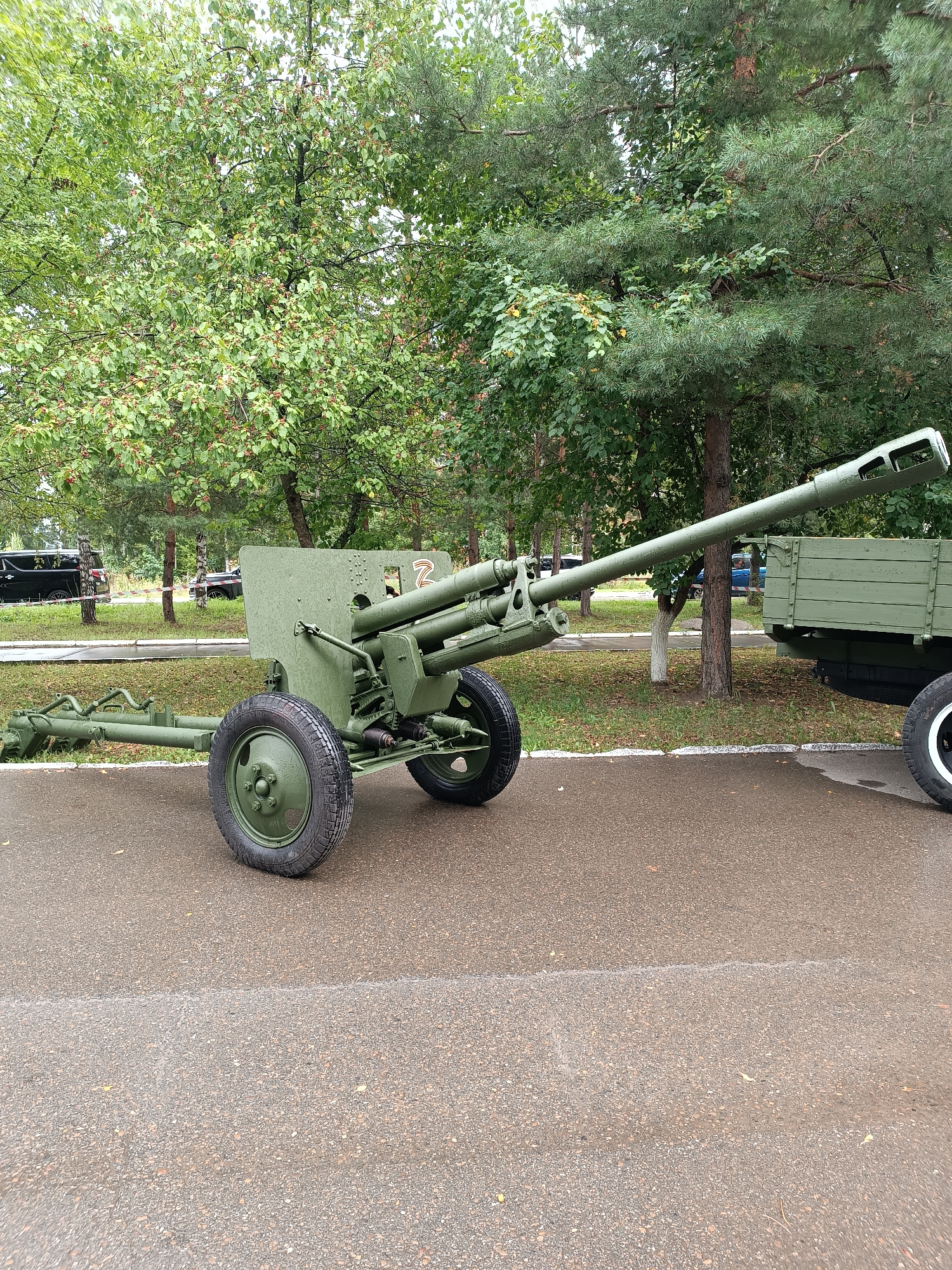 An artillery piece is parked on a paved surface, likely a street or pathway, in an outdoor setting. The artillery piece is painted olive green and has two wheels with black tires. It is positioned at an angle, with its barrel pointing to the right. Behind the artillery piece, a green wooden trailer is visible. The background consists of trees with green foliage. The scene appears to be in Chaikovsky, Russia, as suggested by the context of the provided information. There are no people visible in the image.