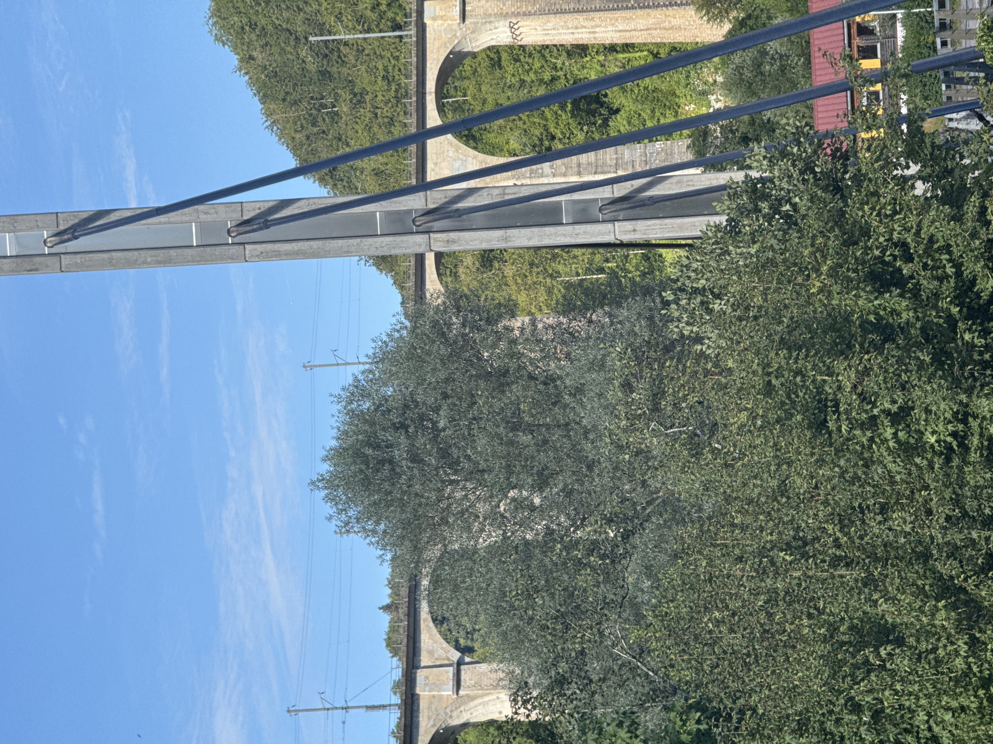 The image depicts a rural landscape in Clos du Doubs, Switzerland, featuring a large, arched stone railway viaduct in the background, partially obscured by dense green foliage. The viaduct, constructed of light-colored stone, has multiple arches spanning across a valley. Above the viaduct, power lines and railway tracks are visible. In the foreground, a tall, modern concrete pillar with dark cables attached stands prominently, suggesting it is part of a newer infrastructure project that may be integrated with or adjacent to the historic viaduct. The pillar is segmented and has a vertical light panel in its center. A lush, green hillside covered in trees rises behind the viaduct, indicating a natural, forested environment. In the lower right quadrant of the image, a building with a red roof and yellow siding is partially visible, hinting at a nearby settlement or industrial area. The sky is a clear, bright blue with scattered wispy white clouds, indicating fair weather. The overall scene captures a juxtaposition of historical and modern infrastructure within a natural Swiss setting.