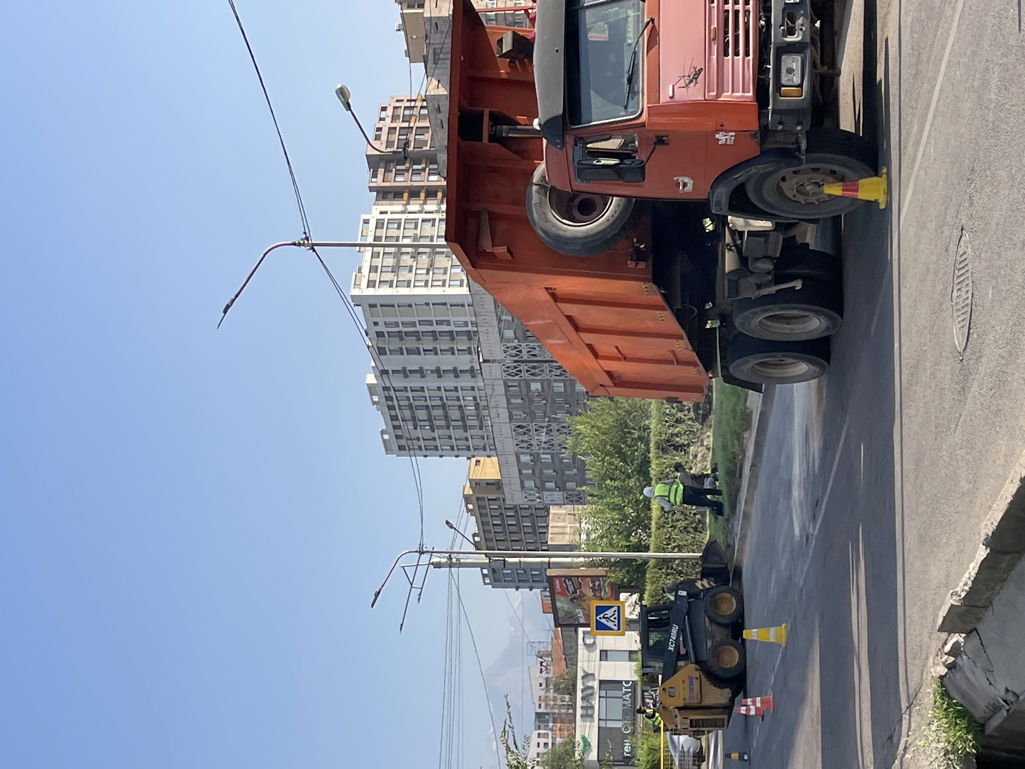 This image depicts a street scene in Almaty, Kazakhstan, likely during daytime construction work. In the foreground, a large orange dump truck with its bed raised is parked on the right side of the frame. Adjacent to the truck, on the road, are traffic cones, including a yellow and red one. To the left of the dump truck, a compact yellow construction vehicle, identified by the text "XC740RU", is positioned. Several workers in high-visibility vests are present. One worker stands near the compact vehicle, while another, wearing a white hard hat and a yellow vest, is visible further back, appearing to be engaged in work on the ground. The background reveals a cityscape with several multi-story buildings of varying architectural styles. A street lamp is visible in the middle ground, and power lines are strung across the sky. The sky is clear and blue, suggesting fair weather. A sign with a yellow diamond shape and a black symbol is visible on a pole. The overall impression is one of an active construction or road repair site within an urban environment.