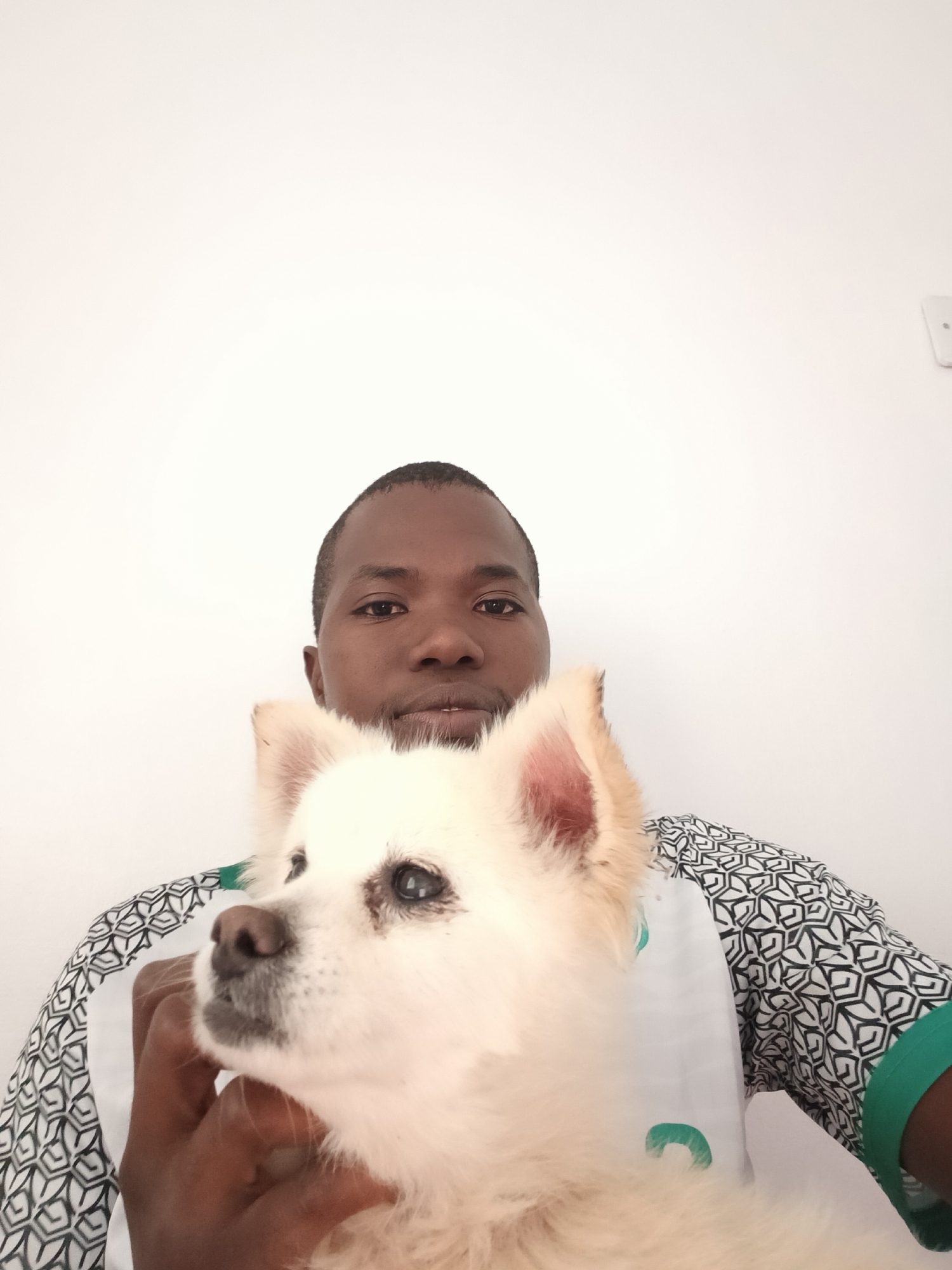 A close-up, portrait-style image features an adult male holding a small, cream-colored, fluffy dog against a plain white background. The male, with a dark complexion and short, dark hair, looks directly at the camera with a neutral expression. He wears a collared shirt with a black geometric pattern on white fabric on the sleeves and shoulder, a white collar, and a visible green trim on the right sleeve cuff. His left hand is shown holding the dog. The dog is positioned in front of the male's chest and lower face, its head turned slightly to its left, away from the camera. It has pointed ears, a dark nose, and dark eyes with minor tear staining. The environment is an indoor setting with uniform lighting. The specific location within Nairobi, Kenya, is not discernible from the visual content.