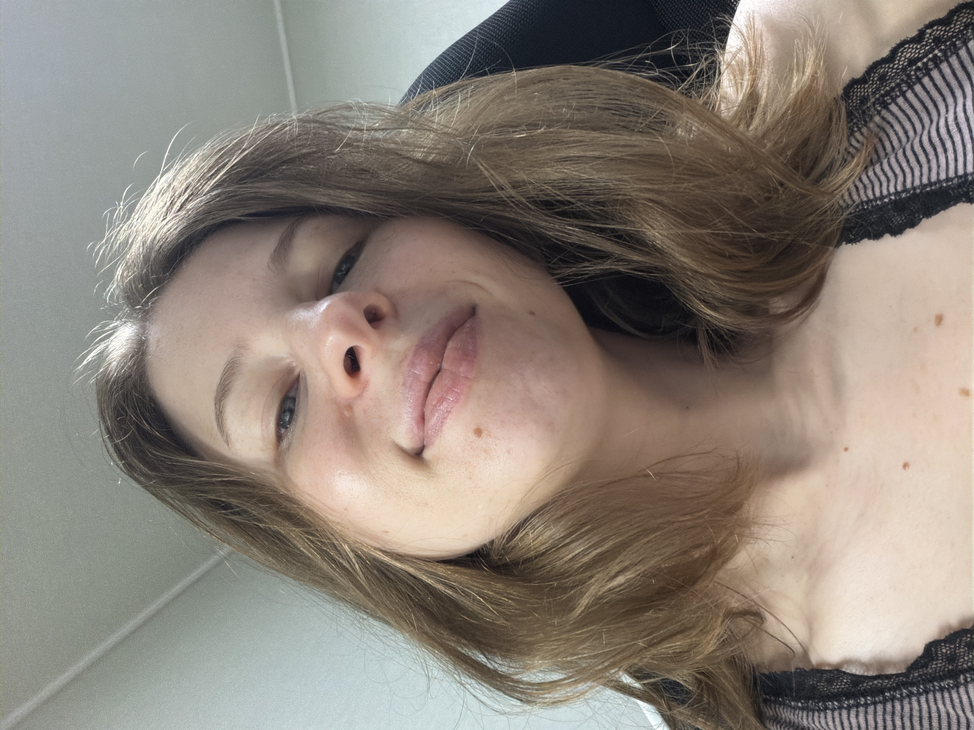 The image is a close-up, low-angle selfie of a young woman with light brown hair and fair skin. She is looking towards the camera with a slight smile. Her hair is shoulder-length and appears somewhat tousled. She is wearing a striped top with black lace trim. The background is blurred, but appears to be an indoor setting with a ceiling visible at the top. The lighting suggests an indoor environment, possibly a room. There are no discernible landmarks or features that would indicate a specific location in Dongducheon-si, South Korea.