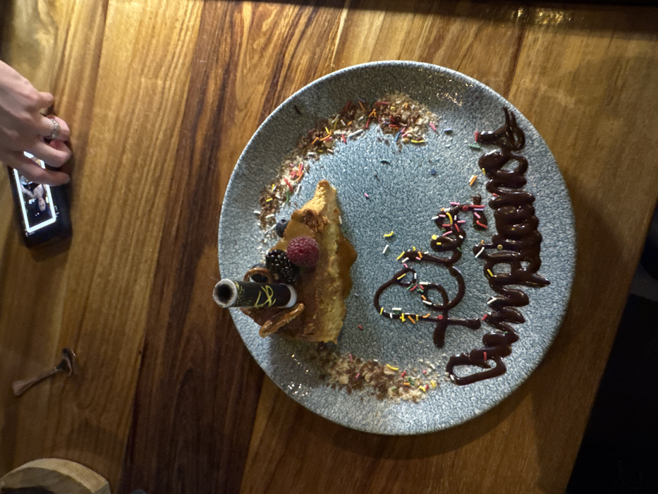 A slice of cake adorned with berries and a decorative cylinder sits on a speckled blue plate. Chocolate syrup spells "Happy Birthday" amidst colorful sprinkles. The plate rests on a wooden table. A hand holding a phone, a small spoon, and a portion of a wooden object are visible on the side.