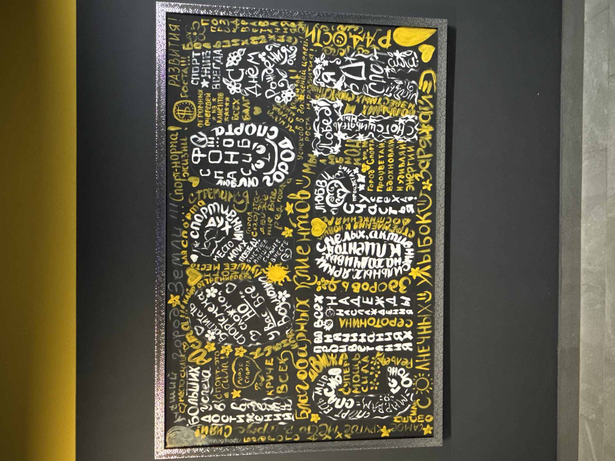 The image shows a framed print hanging on a wall, likely indoors. The print is predominantly black with white and yellow lettering in a densely packed, almost chaotic arrangement. The lettering appears to be in Russian, with a variety of words and phrases related to sports, health, success, and positive affirmations. The print is surrounded by a metallic, possibly silver frame. The wall the print is hanging on is a dark gray, and there is a portion of a mustard-yellow wall visible on the left side of the frame and a light-gray wall to the right of the frame. Given the context and text content, it likely represents artwork displayed in a sports facility, gym, or motivational space. The location is possibly Votkinsk, Russia, as it is implied by the Russian text.