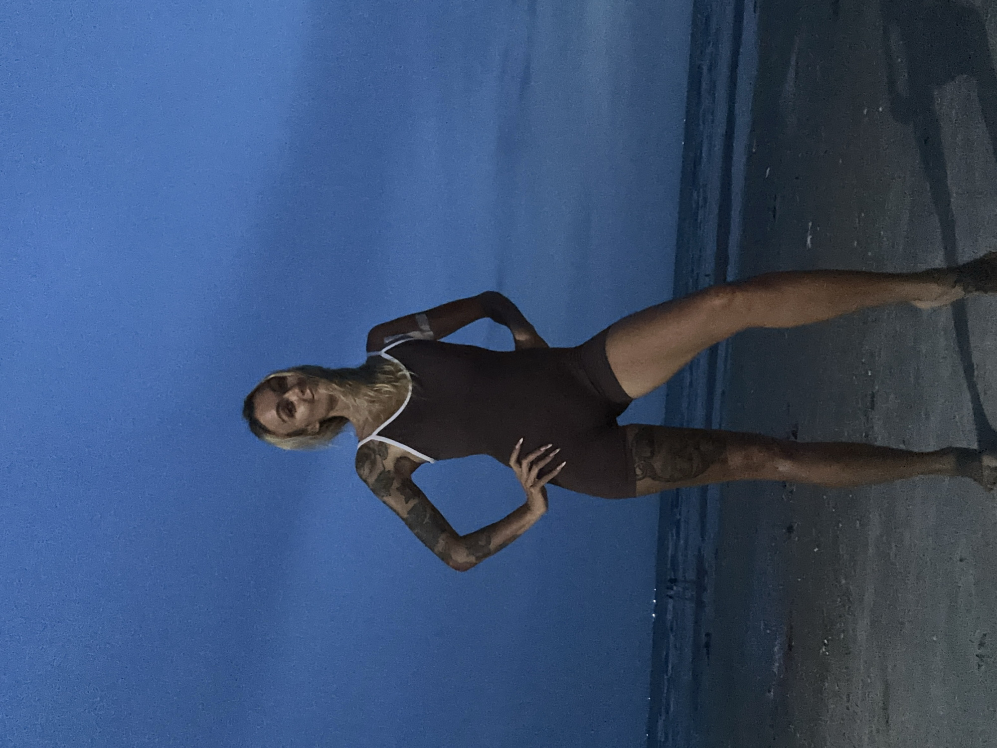 A woman with blonde hair and extensive tattoos stands on a beach at dusk. She is wearing a dark one-piece swimsuit with white trim. Her hands are on her hips. The ocean is visible behind her, with faint lights in the distance. The sky is a deep blue. The image appears to be taken in Mũi Né, Vietnam.