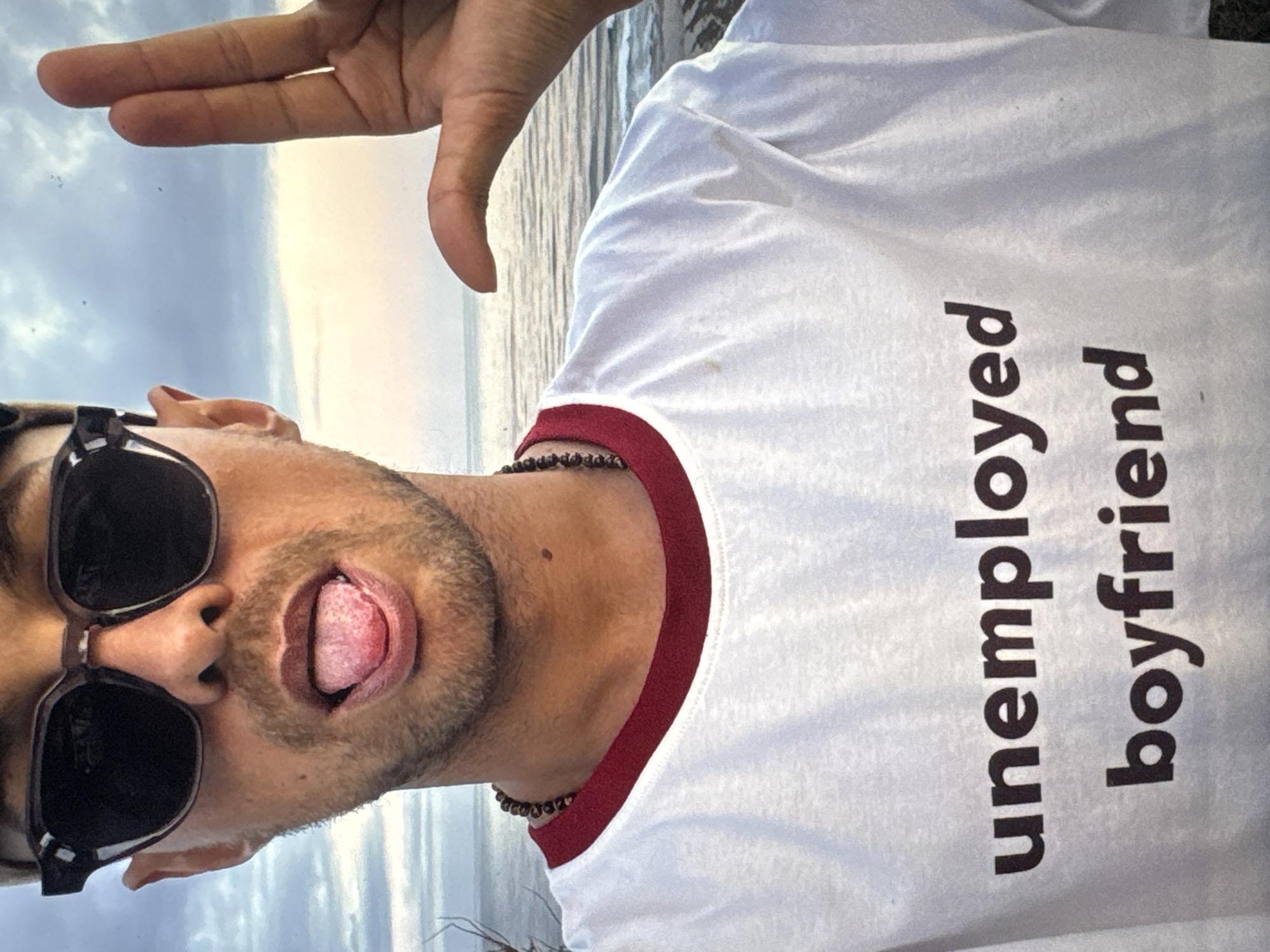 The media file is a close-up, self-portrait photograph of a young man with sunglasses and a white t-shirt that has "unemployed boyfriend" printed on it. The man is sticking his tongue out, and his hand is raised in the frame, obscuring part of the sky. The background suggests an outdoor setting with a cloudy sky. The lighting indicates it is likely daytime. The man appears to be playful or making a humorous gesture. The location context provided is Kuta Selatan, Indonesia.