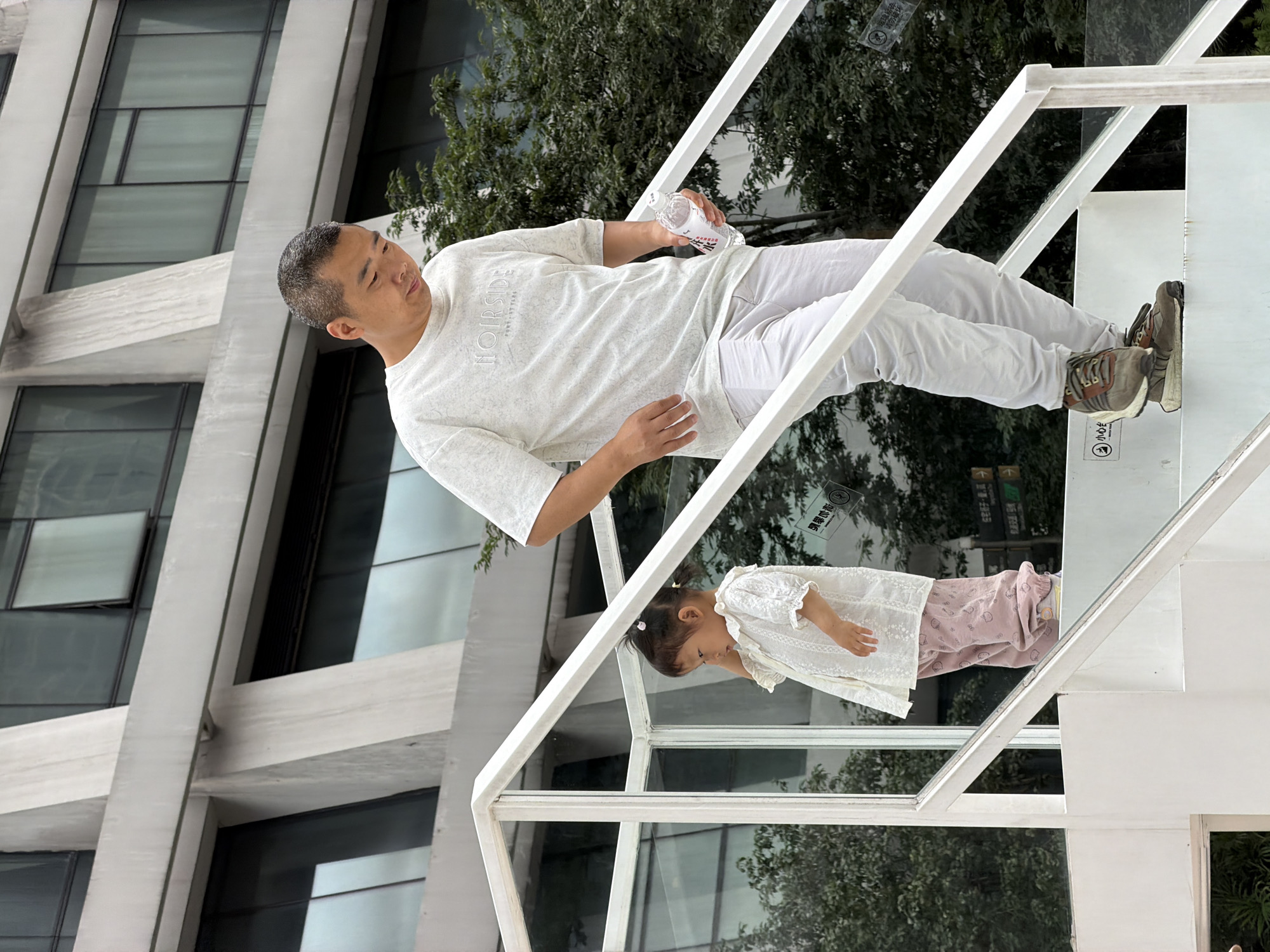 In Yuzhong District, China, an adult male wearing a light-colored t-shirt and white pants is walking down white stairs. He is holding a clear plastic bottle of water in his right hand. To his left and slightly behind him, a young child, appearing to be a girl, is standing on the stairs. She is wearing a white lace top and pink patterned pants. The background features a modern building with glass windows and concrete structures, along with greenery from trees. The stairs have white railings and glass panels.