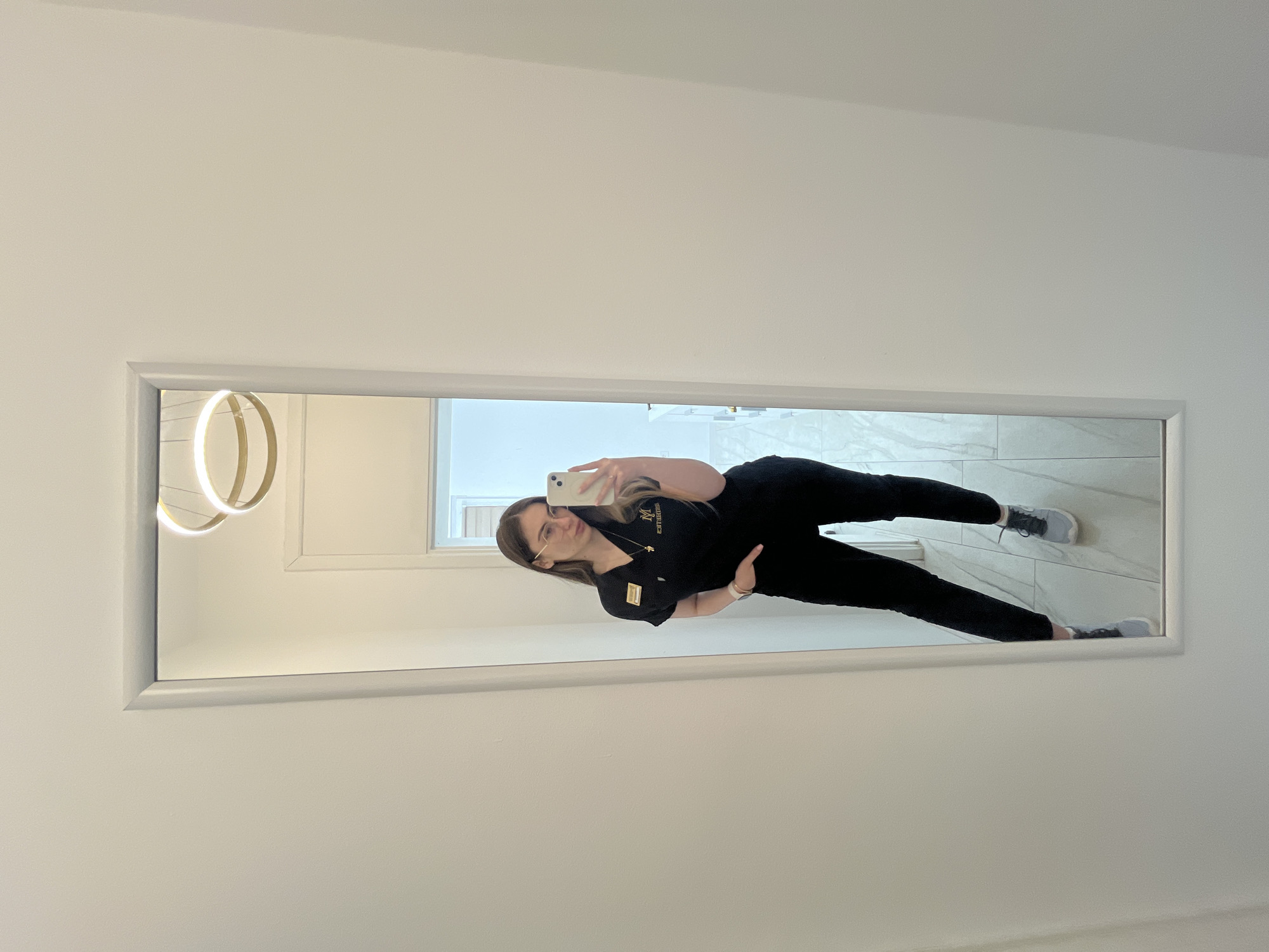 A full-length mirror is mounted on a white wall in a room. The reflection in the mirror shows a woman wearing black attire and grey sneakers taking a selfie with her phone. The floor is tiled. The room has a modern aesthetic, as indicated by the circular light fixture on the left side of the mirror.