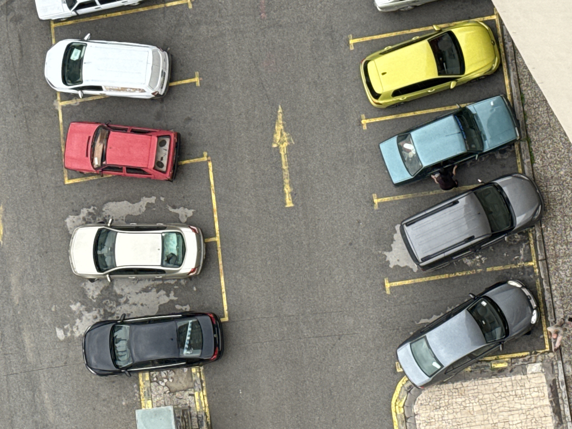 An aerial view shows a parking lot with several cars. In the top row, from left to right, are a black sedan, a silver sedan, a red sedan, and a white sedan. In the bottom row, from left to right, are a dark gray sedan, another dark gray sedan, a light blue sedan, and a yellow sedan. A person is partially visible near the light blue sedan, leaning towards it. The parking lot is paved with asphalt and marked with yellow lines. There are some areas of wetness or staining on the asphalt. The scene is set in the city of Konya, Turkey, as indicated by the context.