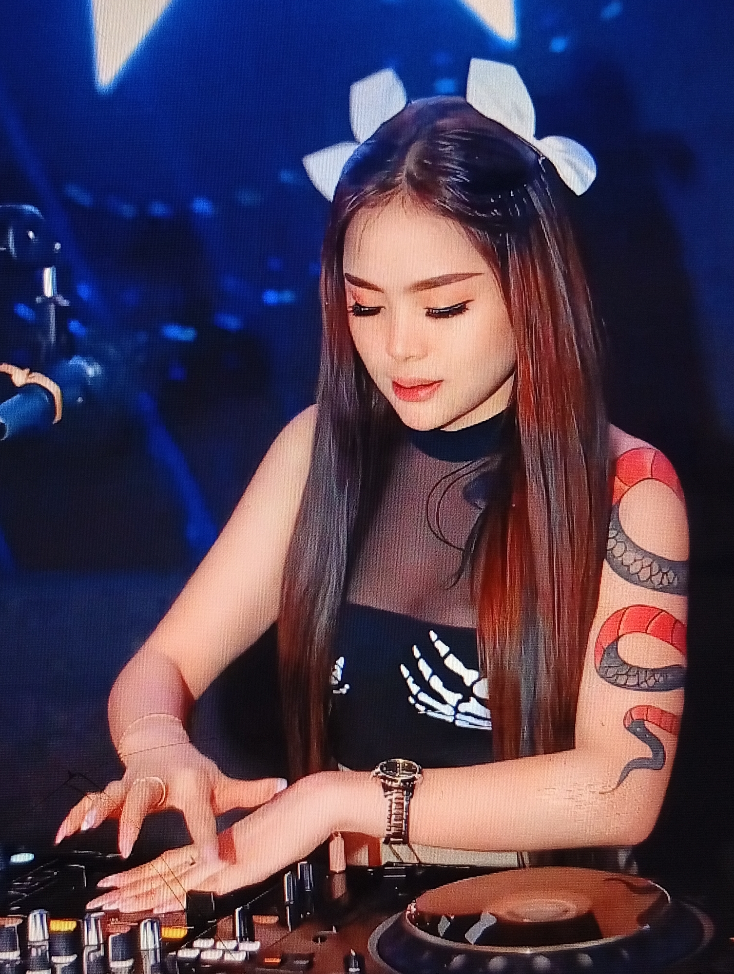 A young woman with long, reddish-brown hair and white bow accessories in her hair is standing and operating a DJ mixer. She is wearing a black, sleeveless top with a sheer neckline and a graphic of a skeleton hand. A large tattoo of a coiled snake with red and black segments covers her right arm. She is wearing a watch on her left wrist and multiple rings on her fingers, which are positioned over the DJ controls. The background is dark with blurred blue and white lights, suggesting a performance or club setting. The image is a close-up shot focused on the woman and the DJ equipment. There is no specific indication of the location being in Lhokseumawe, Indonesia.
