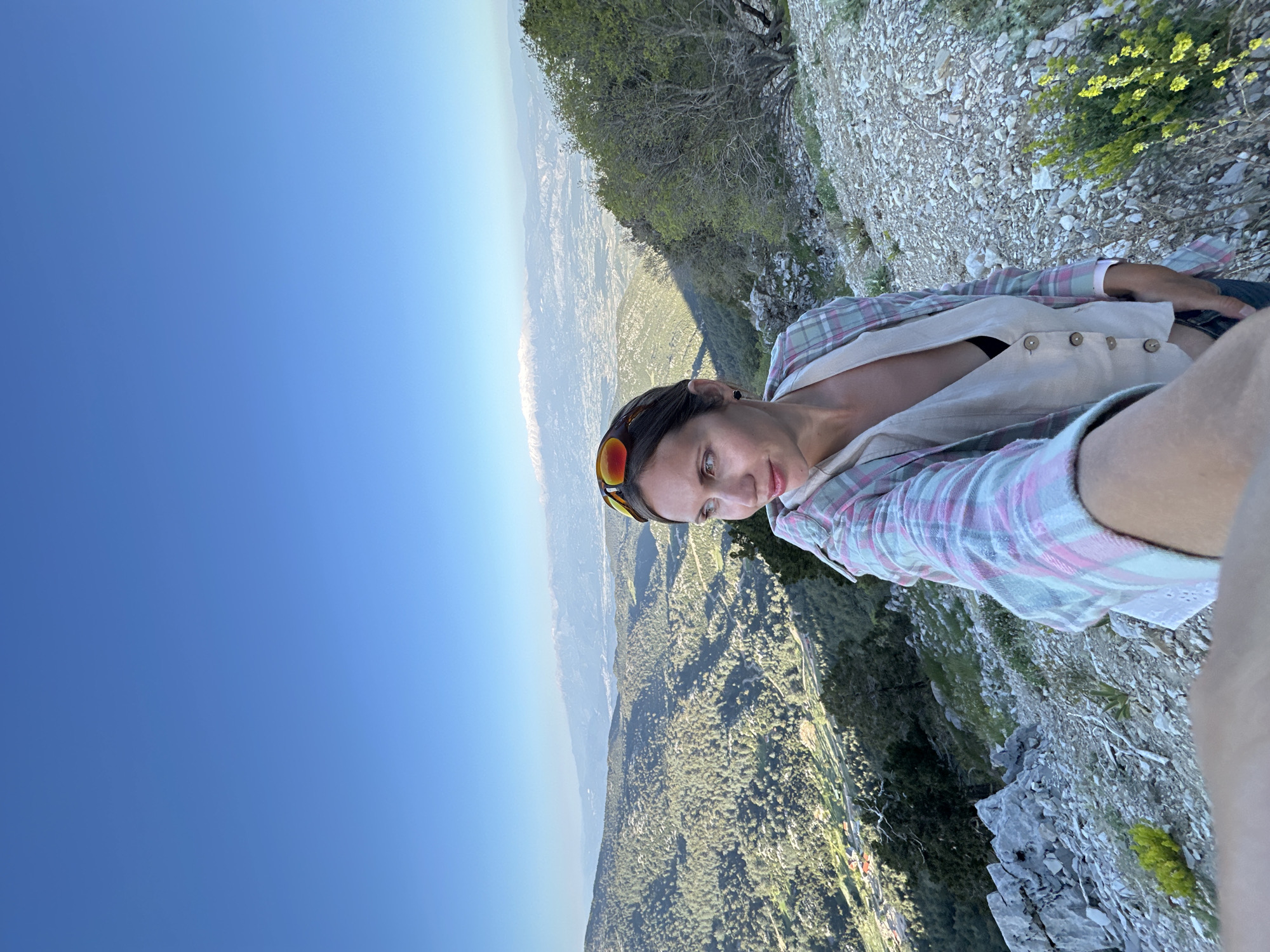 A woman stands on a rocky cliff overlooking a valley, potentially near Fethiye, Turkey, judging by the terrain. She is wearing sunglasses, a plaid shirt, and a light-colored vest. The background shows a landscape with vegetation and settlements. The sky is blue. The image appears to be a selfie.